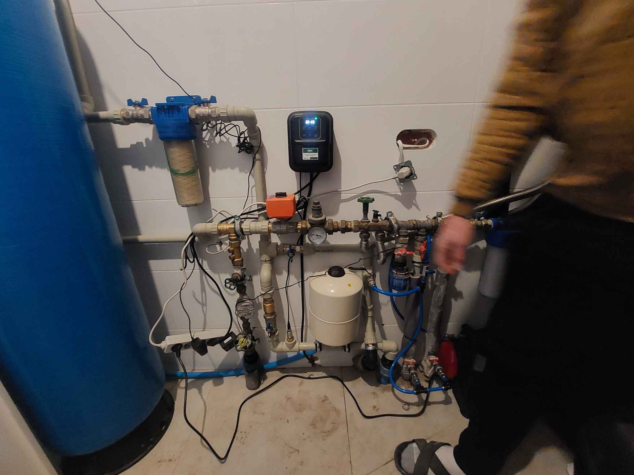 Kompleksowa instalacja wodna z filtrem, zbiornikiem hydroforowym i sterownikiem elektronicznym na tle białych płytek, widok z boku z fragmentem osoby.