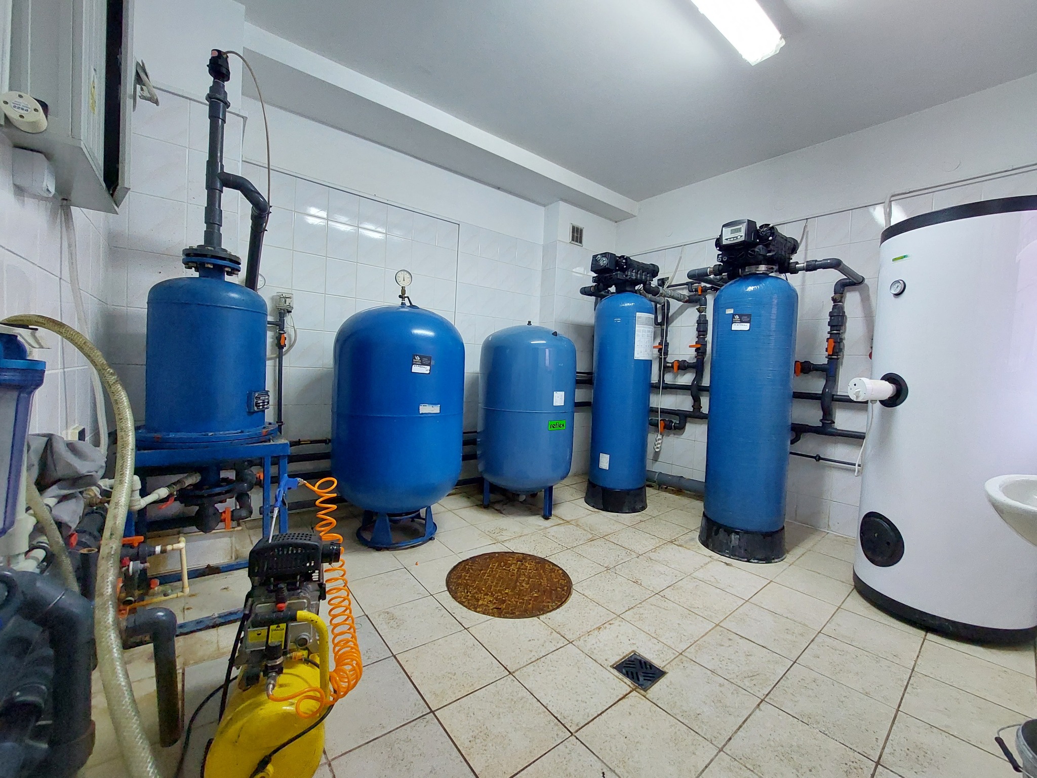 Pomieszczenie techniczne z niebieskimi zbiornikami ciśnieniowymi, filtrami do wody i żółtym kompresorem, widoczna studzienka rewizyjna w podłodze z płytek.