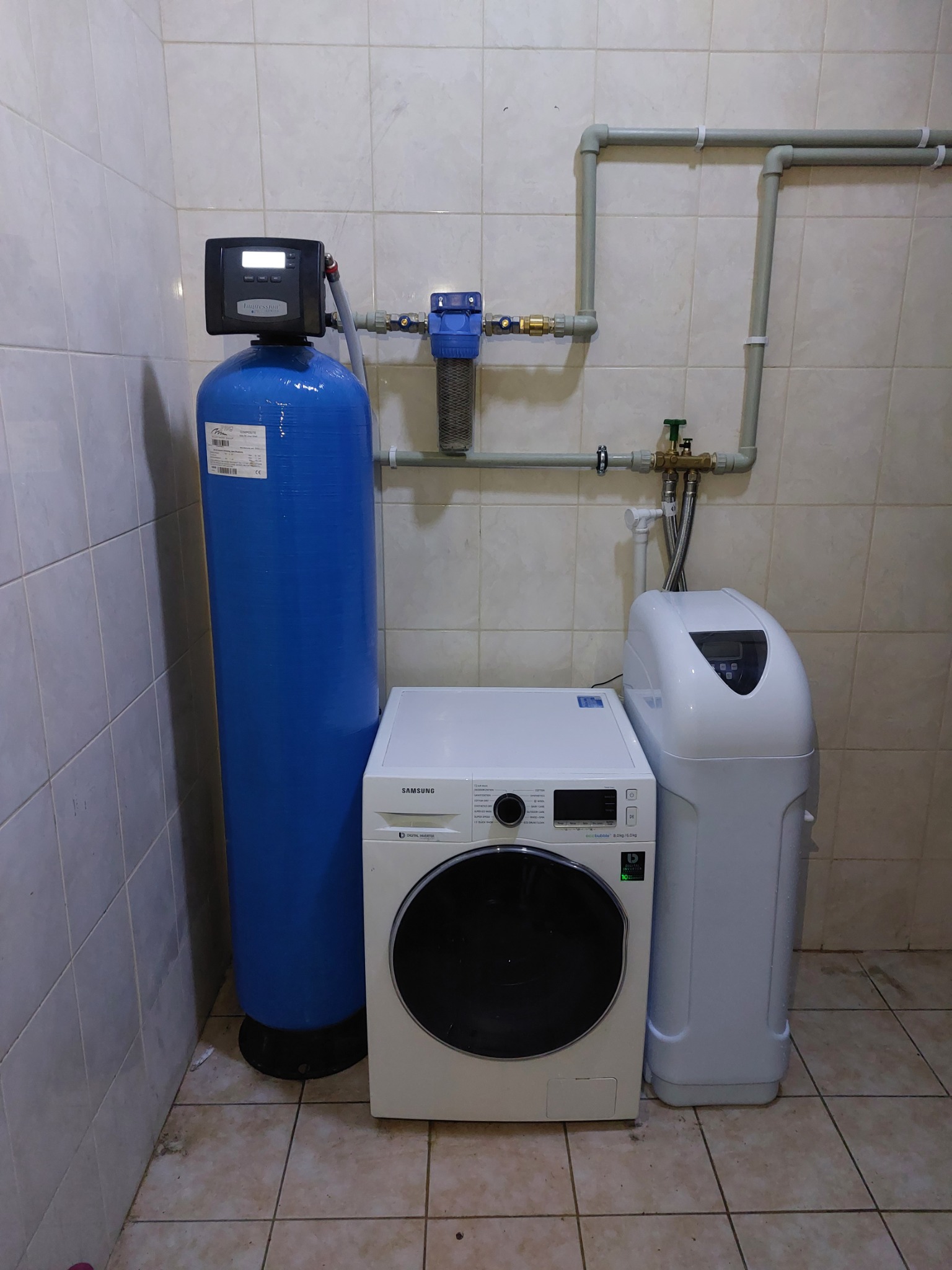 Niebieski zbiornik uzdatniania wody z czarnym sterownikiem, filtr narurowy i biały zmiękczacz wody obok pralki w łazience z beżowymi płytkami.