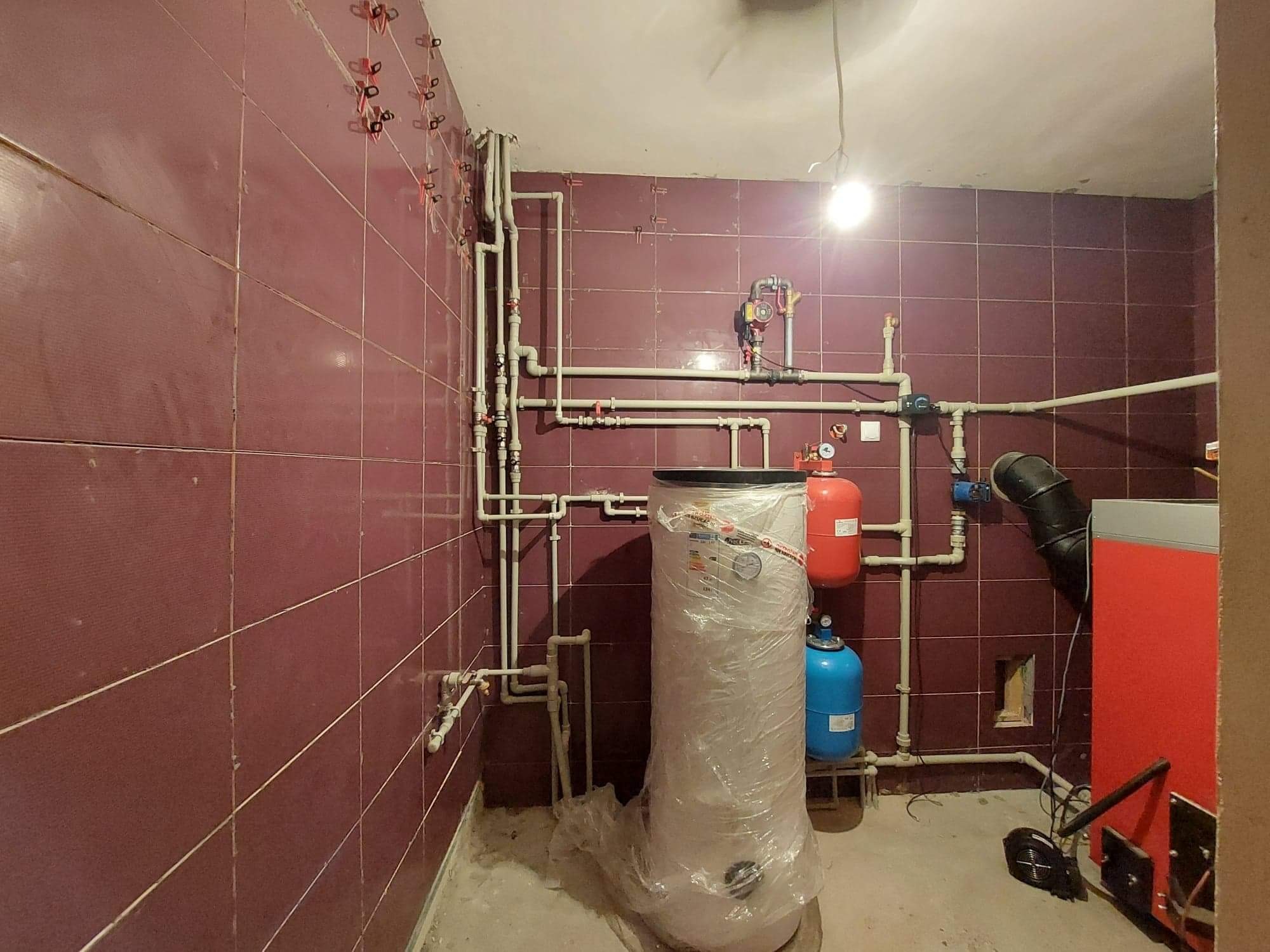 Pomieszczenie z instalacją grzewczą w trakcie montażu, widoczne rury, zbiorniki, piec oraz bojler owinięty folią ochronną na tle bordowych płytek ściennych.