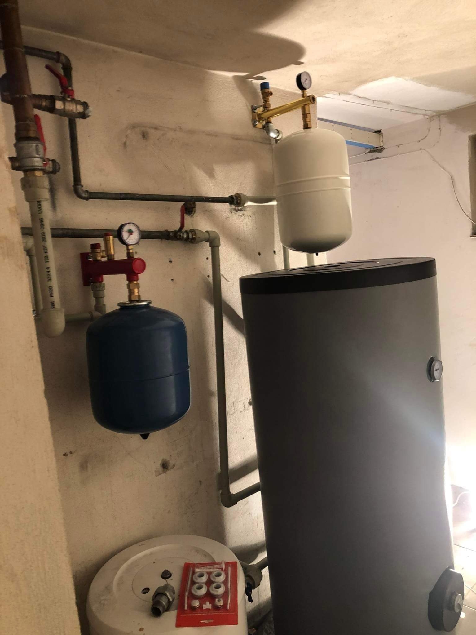 Instalacja grzewcza w piwnicy: Szary zasobnik ciepłej wody z białym naczyniem wzbiorczym u góry, niebieskie naczynie przeponowe, rury miedziane i plastikowe z zaworami, manometry, uszczelki...