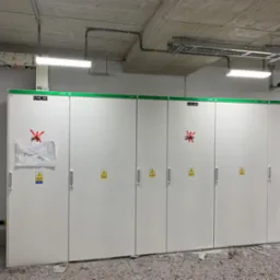 Rozdzielnia elektryczna z białymi szafami, oznaczeniami ostrzegawczymi i zielonymi akcentami, z instalacjami sufitowymi i osłoniętą podłogą.