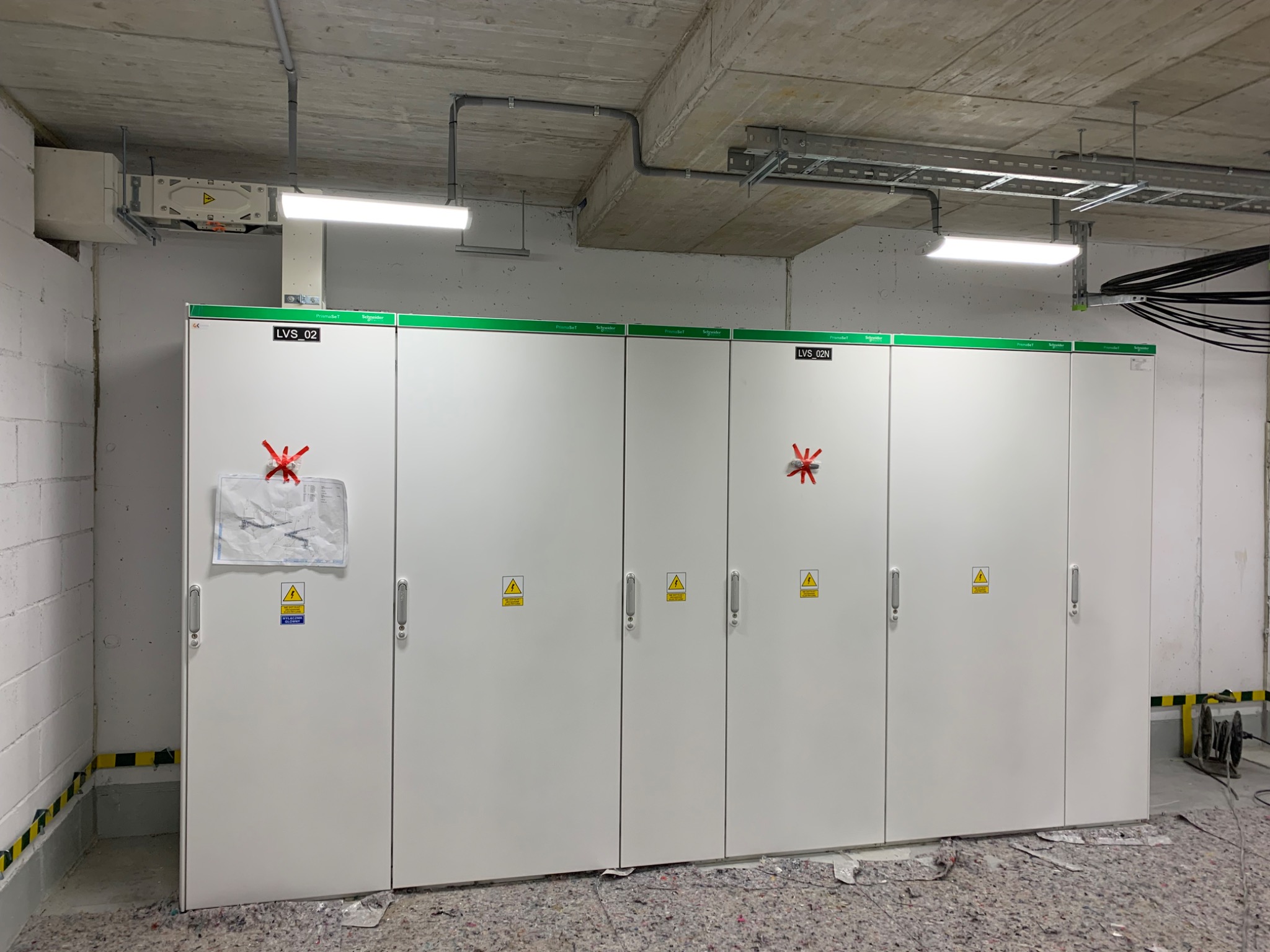 Rozdzielnia elektryczna z białymi szafami, oznaczeniami ostrzegawczymi i zielonymi akcentami, z instalacjami sufitowymi i osłoniętą podłogą.