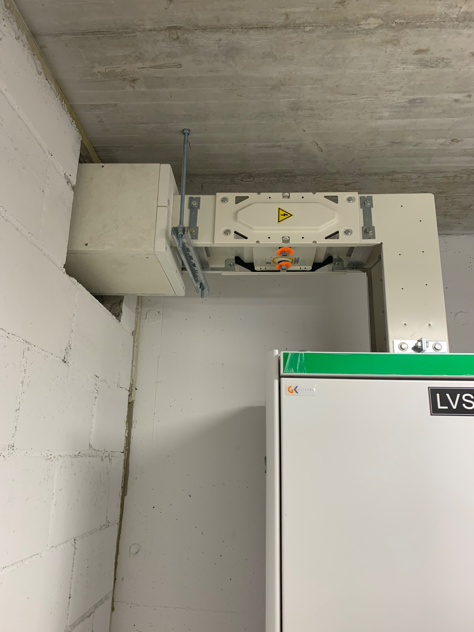 Zamontowana pod sufitem skrzynka elektryczna z symbolem ostrzegawczym wysokiego napięcia, widoczne elementy mocujące, ściana z białej cegły w tle.