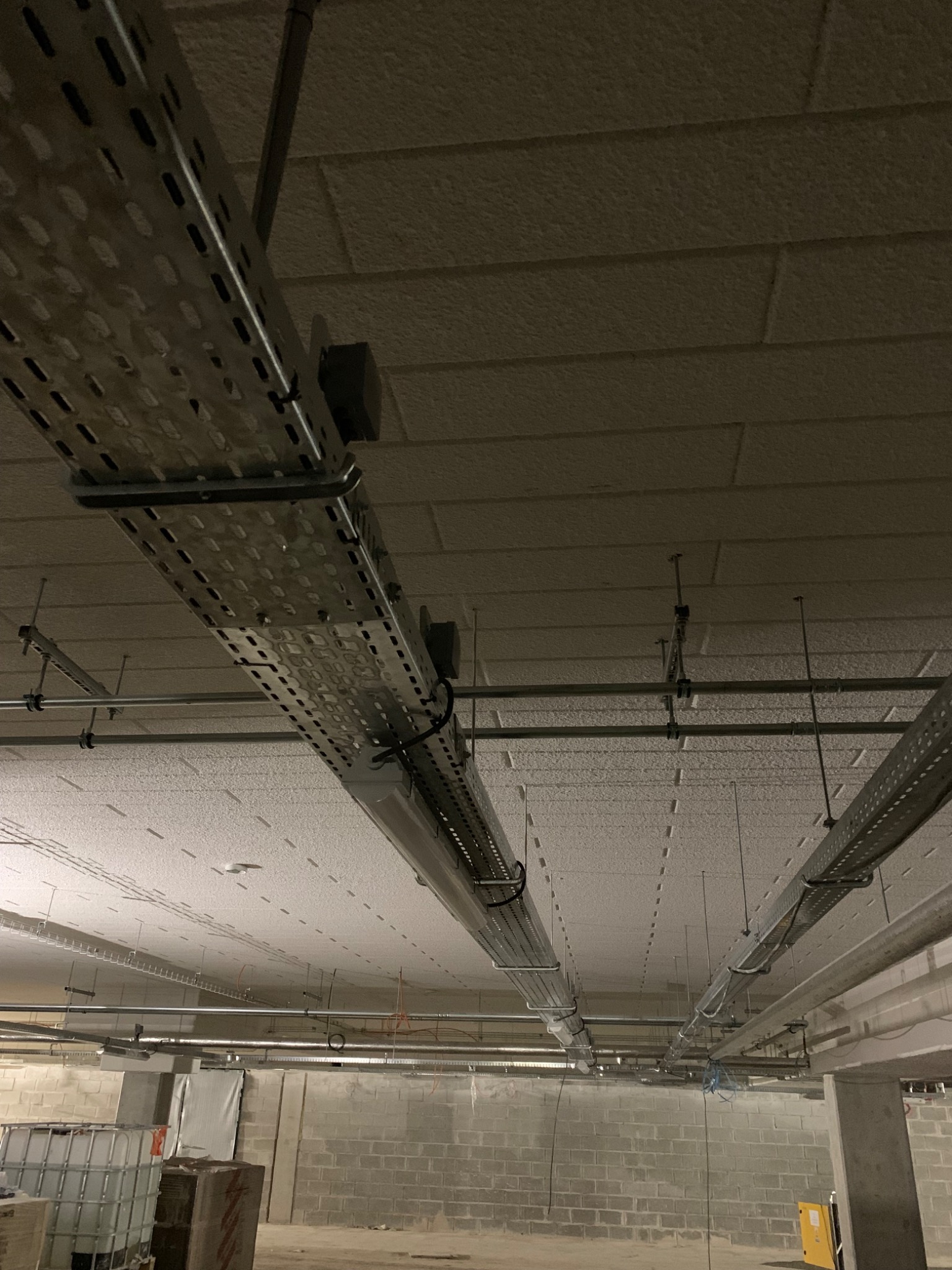 Metalowe korytka kablowe pod sufitem technicznym w surowym wnętrzu, widoczne także rury i instalacje.