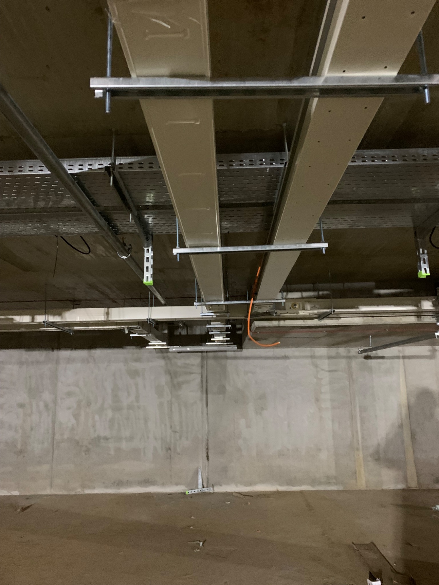 Surowy betonowy sufit z metalowymi korytkami kablowymi i przewodami, widoczne elementy montażowe i poziomica na podłodze.