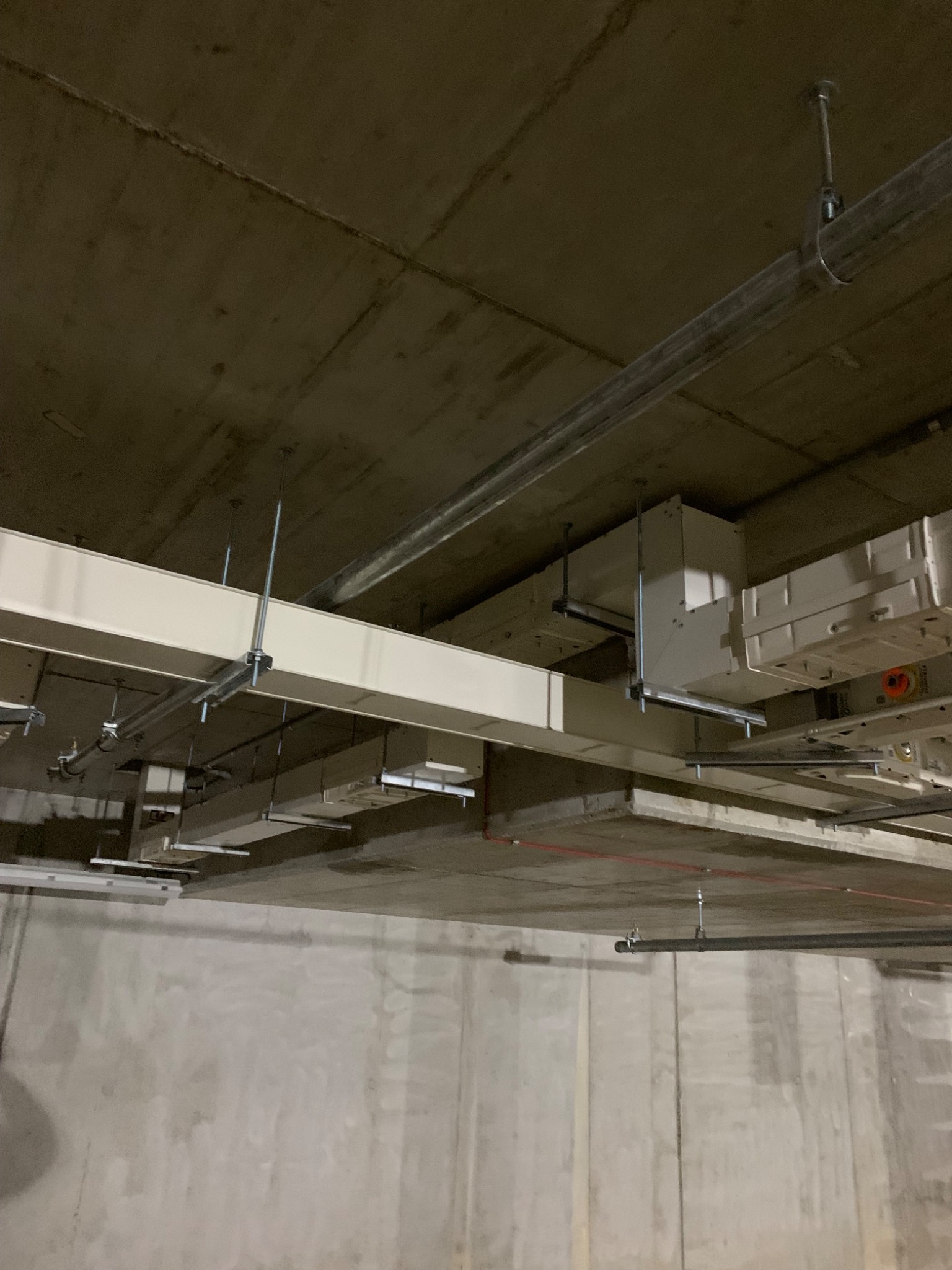 Widok sufitu z zainstalowanymi korytkami kablowymi, rurami i puszkami elektrycznymi, mocowanymi za pomocą metalowych wsporników i śrub do betonowego stropu.