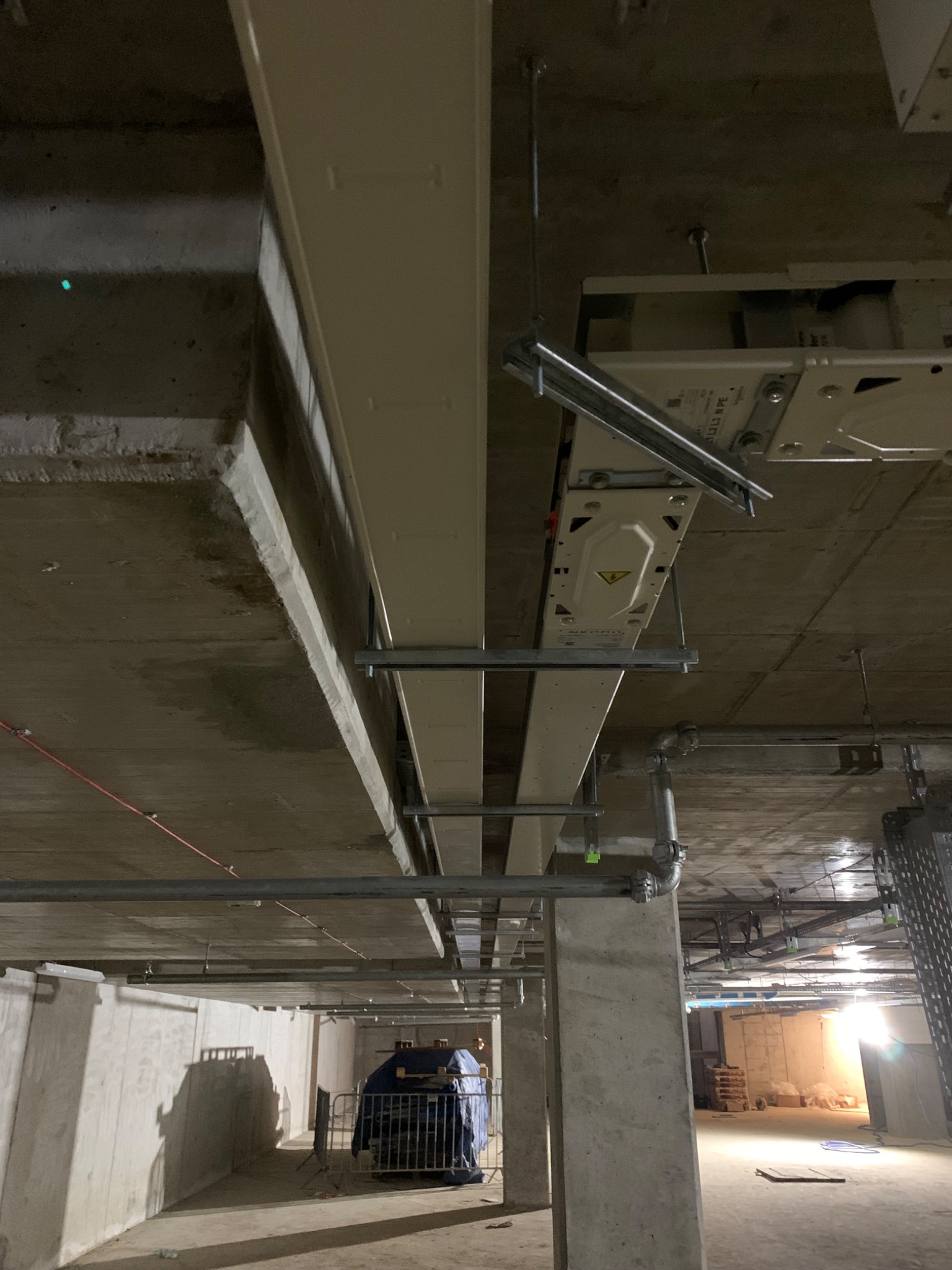 Surowy betonowy strop z zamontowanymi metalowymi konstrukcjami pod instalacje elektryczne i wentylacyjne, widoczne rury i kable, w tle zabezpieczone materiały budowlane.