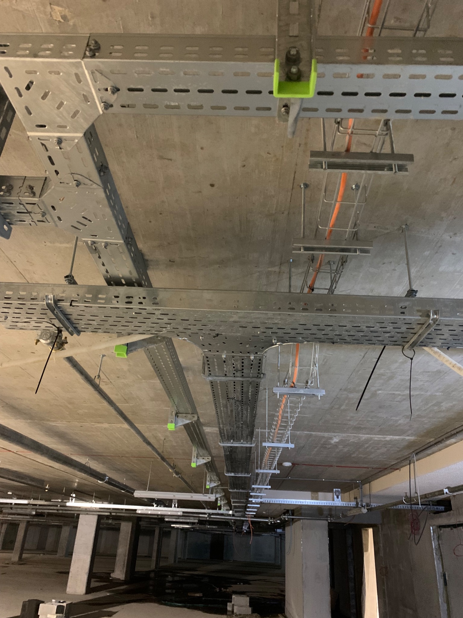 Metalowe korytka kablowe z przewodami zamontowane pod betonowym sufitem w surowym stanie. Widoczne zielone elementy mocujące i różnorodne przewody wewnątrz korytek.