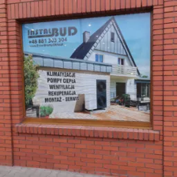 Reklama firmy InstalBud na oknie budynku z cegły, prezentująca dom z pompą ciepła, wentylacją i klimatyzacją.