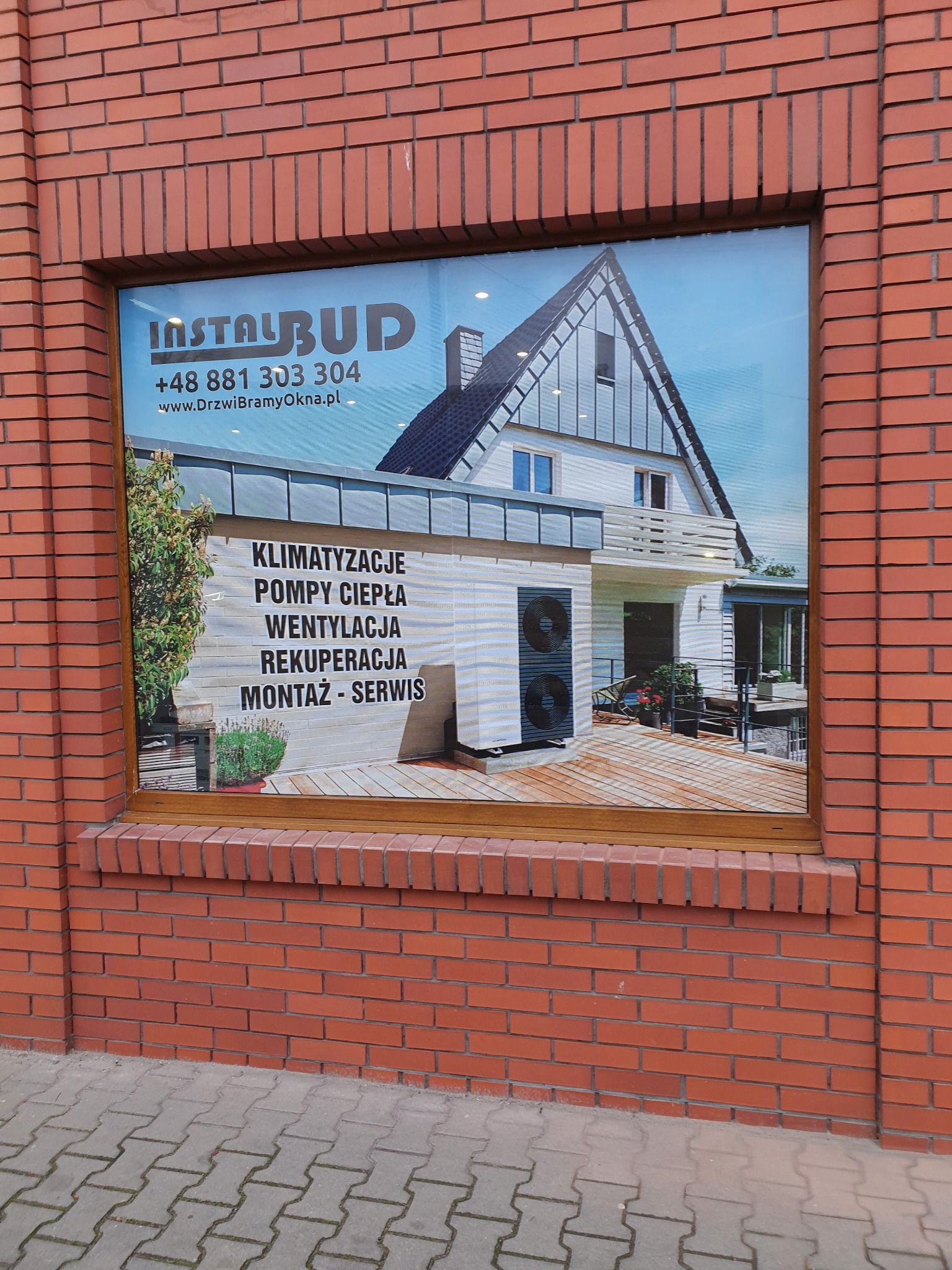 Reklama firmy InstalBud na oknie budynku z cegły, prezentująca dom z pompą ciepła, wentylacją i klimatyzacją.