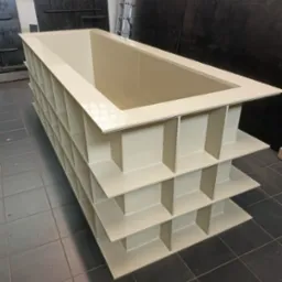 Beżowa konstrukcja stalowa o regularnej strukturze kratownicy, prawdopodobnie element hali, widoczna z perspektywy lekko z góry na tle ciemnej ściany i podłogi z płytek.