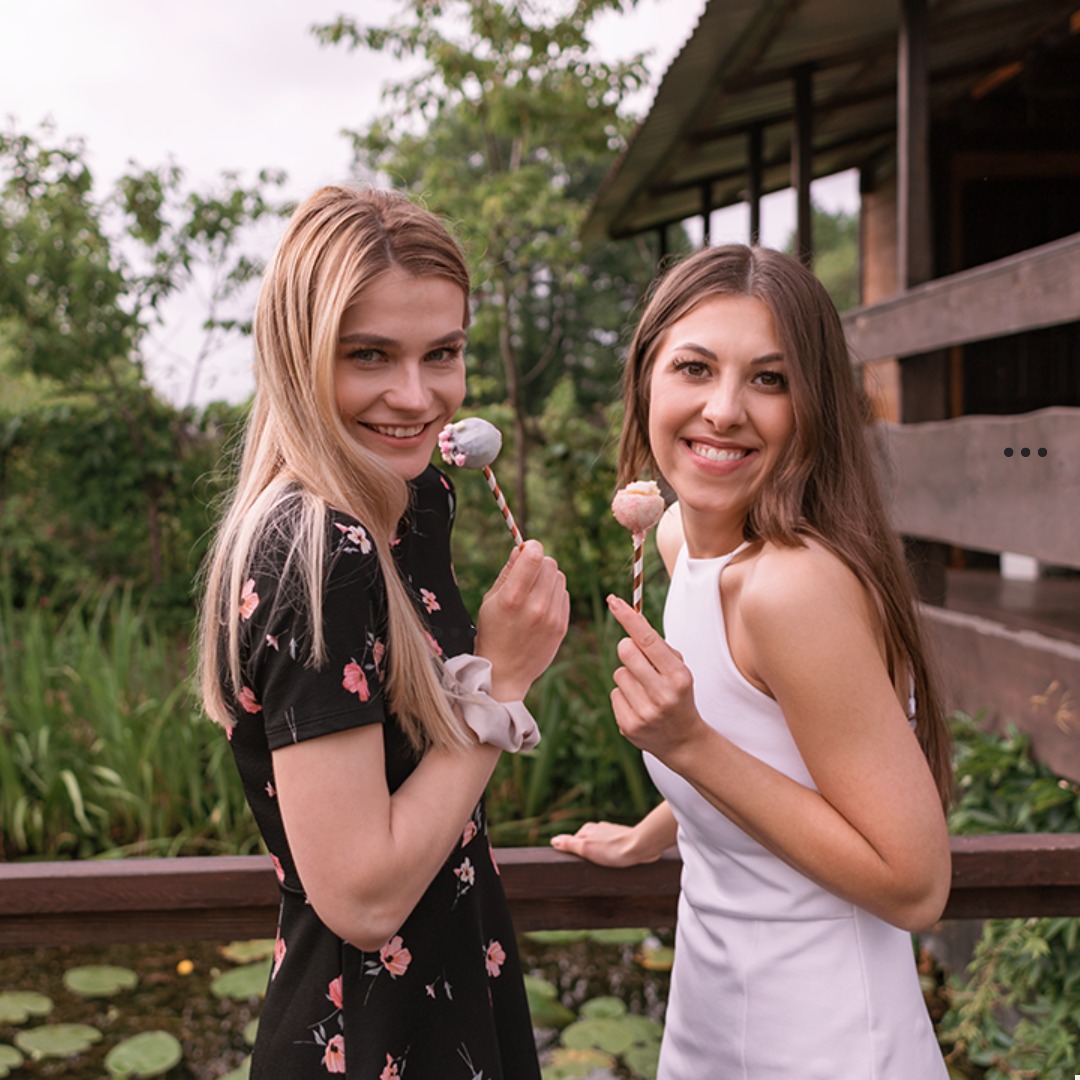 Dwie uśmiechnięte kobiety w Sokołach trzymają lizaki cake pops na drewnianym tarasie z widokiem na staw z liliami wodnymi.