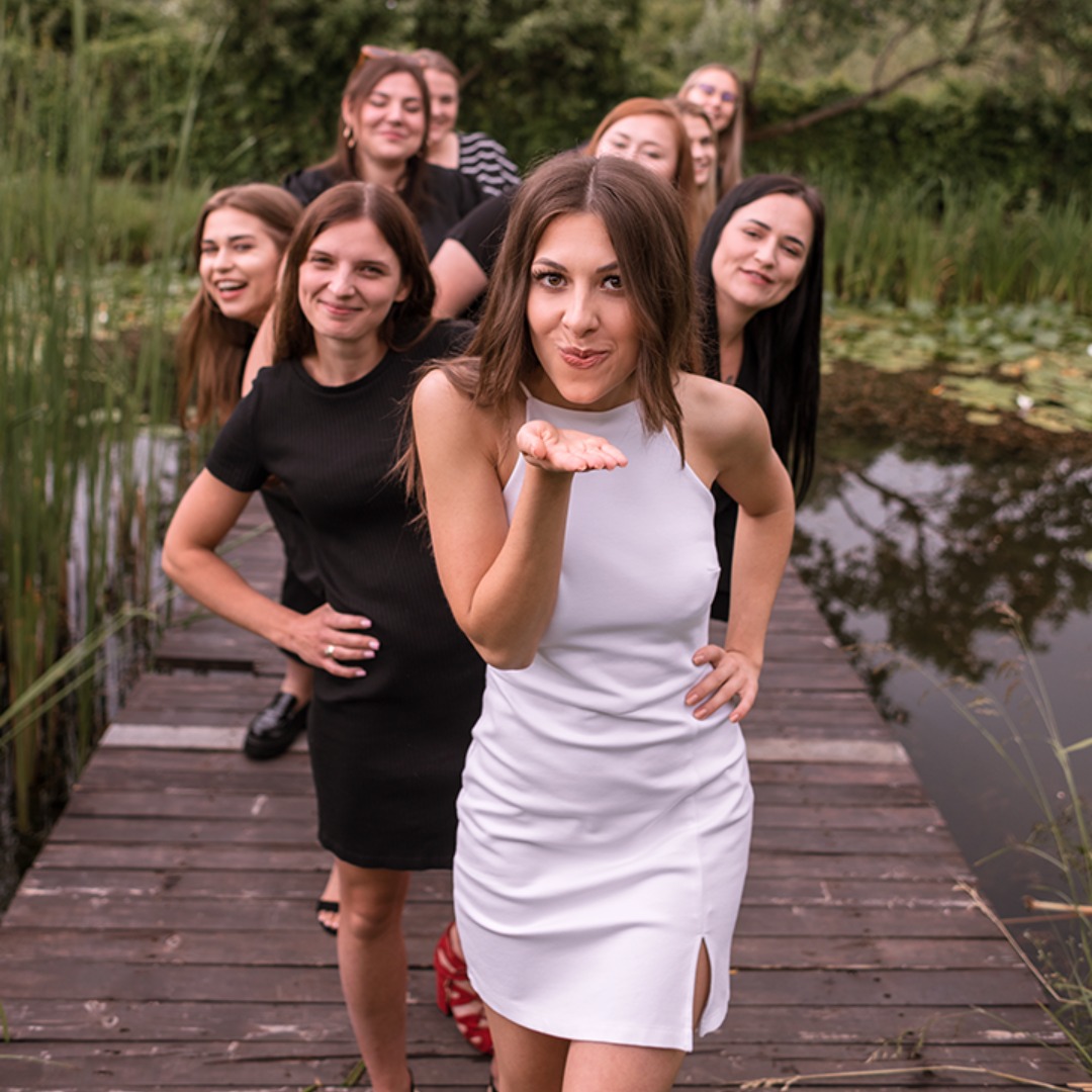 Grupa kobiet w sukienkach pozuje na drewnianym pomoście nad stawem, główna postać w białej sukience wysyła całusa w stronę obiektywu.