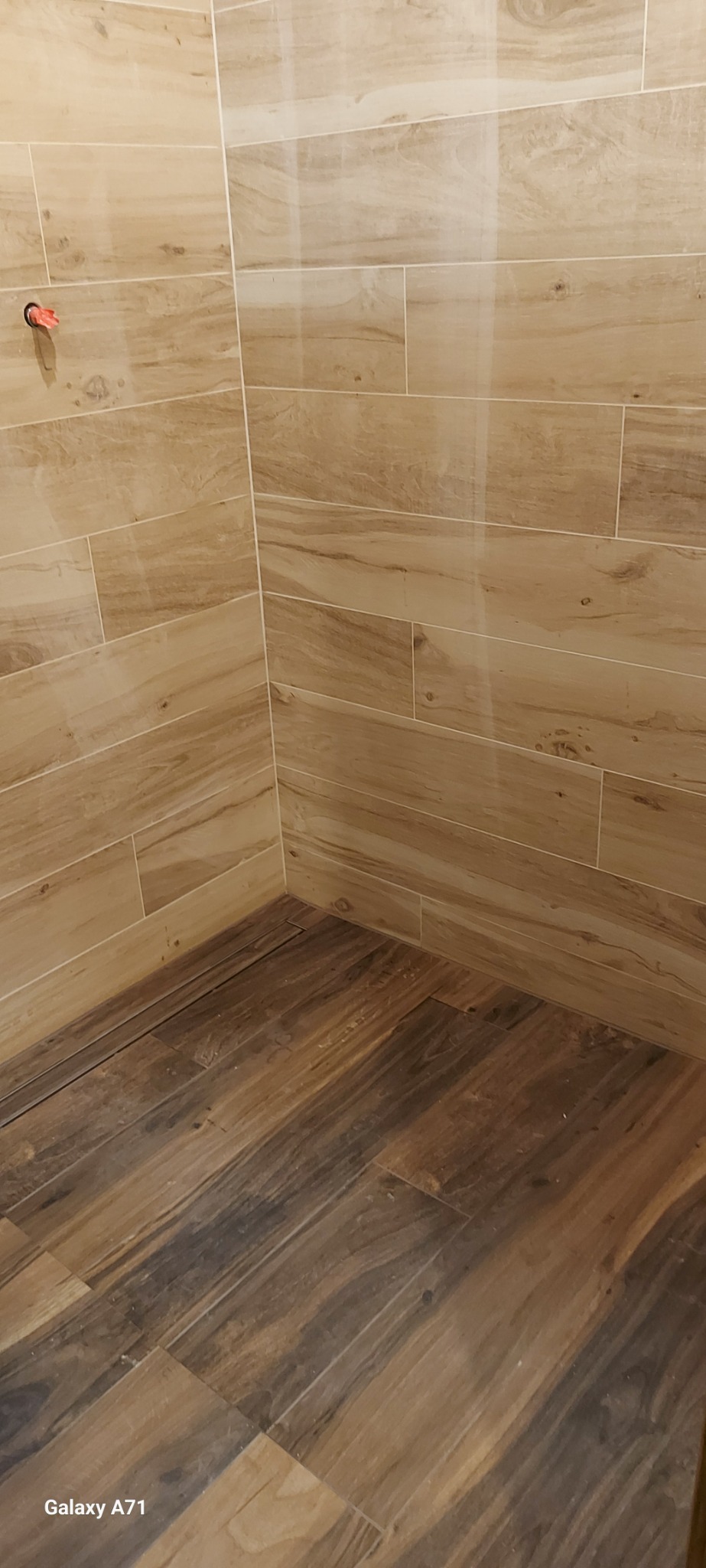 Wykończony narożnik łazienki z beżowymi, podłużnymi płytkami imitującymi drewno na ścianach i ciemnobrązowymi płytkami podłogowymi, również imitującymi drewno.