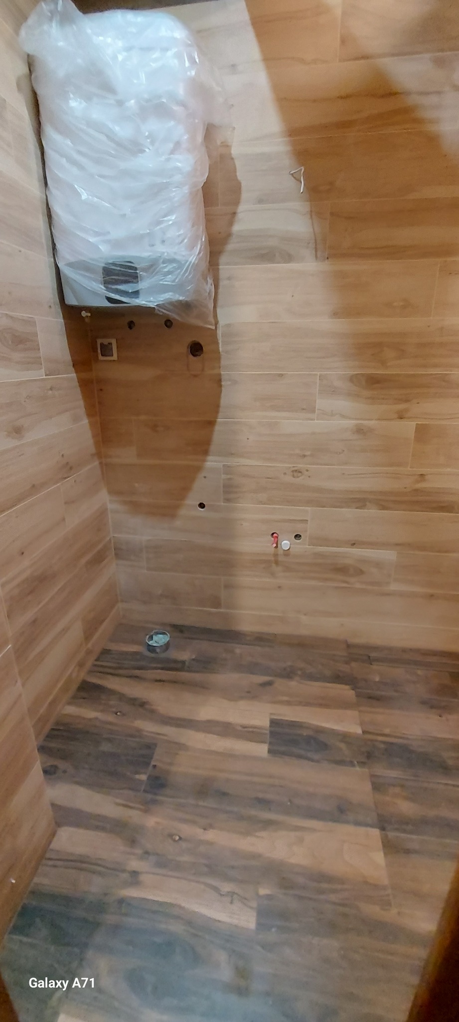 Świeżo wykafelkowana łazienka z drewnopodobnymi płytkami podłogowymi i ściennymi, z zamontowanym, ale jeszcze ofoliowanym bojlerem oraz wyprowadzonymi instalacjami wodno-kanalizacyjnymi.