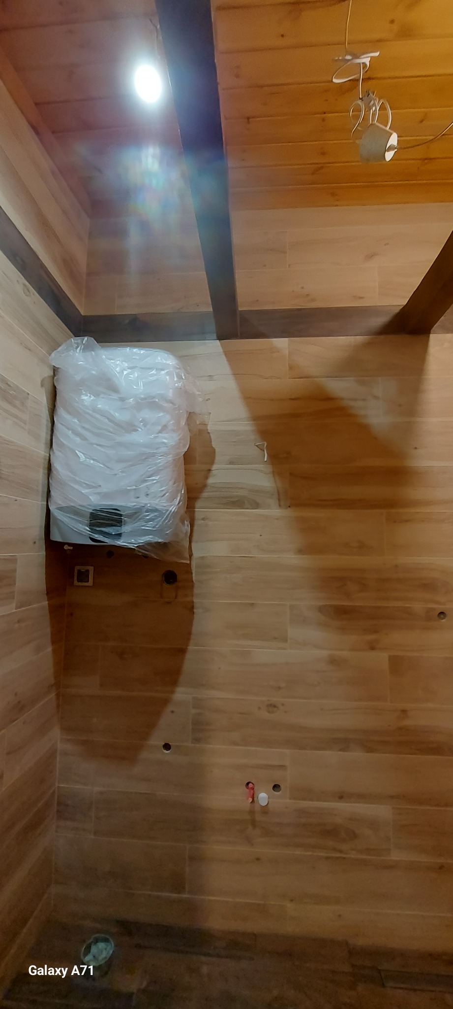 Nowy bojler owinięty folią ochronną, stojący na podłodze wyłożonej płytkami imitującymi drewno w trakcie remontu łazienki.