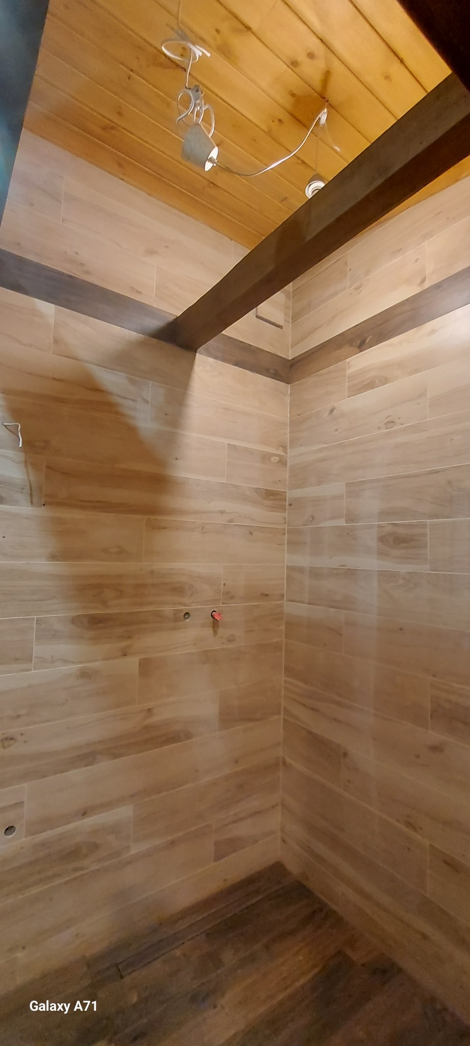 Wykończona łazienka z drewnopodobnymi płytkami na ścianach i podłodze, widoczne otwory na armaturę i drewniana belka pod sufitem z widocznym oświetleniem.
