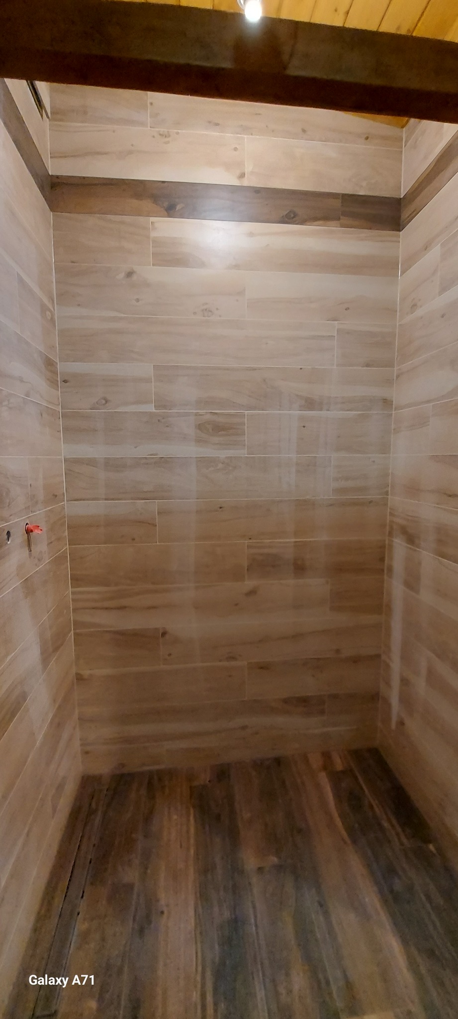 Wnętrze kabiny prysznicowej wykończonej płytkami imitującymi drewno, z widocznymi elementami instalacji wodnej i drewnianą belką stropową u góry.