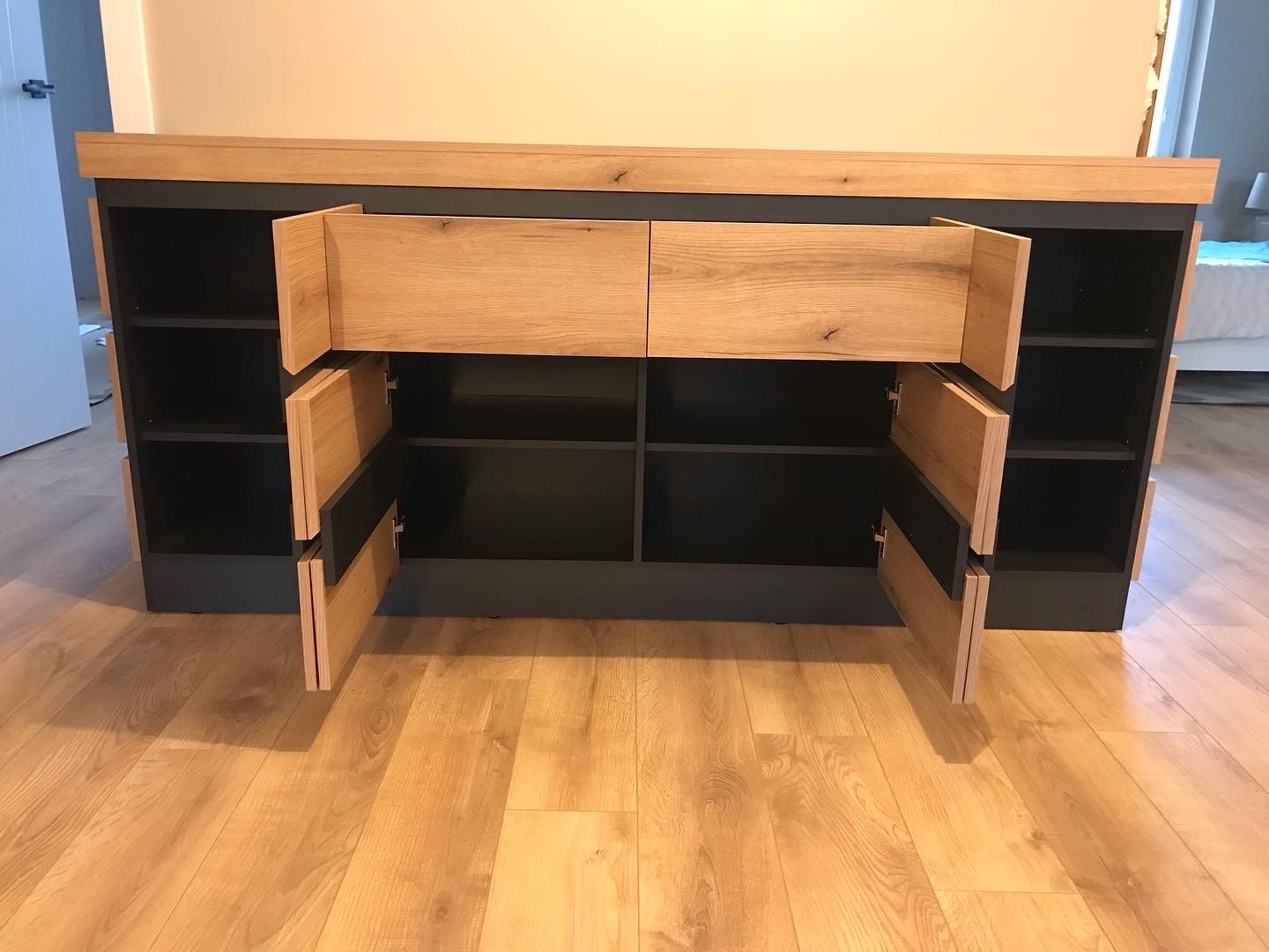 Nowoczesna komoda z otwartymi szafkami i szufladami z jasnego drewna, kontrastująca z czarnym korpusem, ustawiona na drewnianej podłodze.