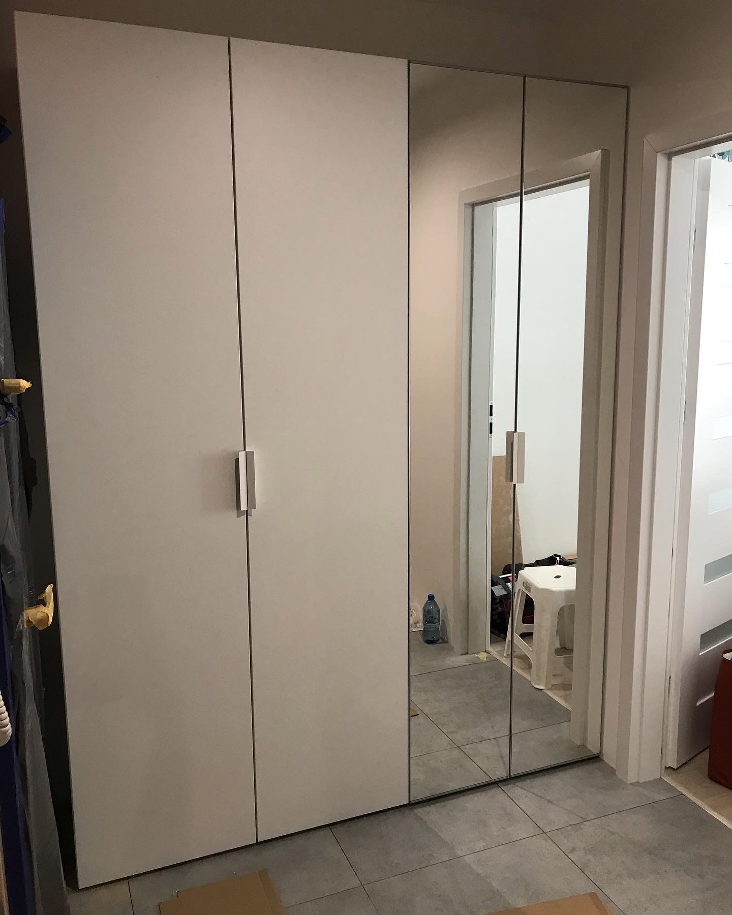 Biała szafa z przesuwnymi drzwiami z lustrem, widoczna częściowo zawartość pokoju za lustrem, w tym białe krzesło i butelka wody.