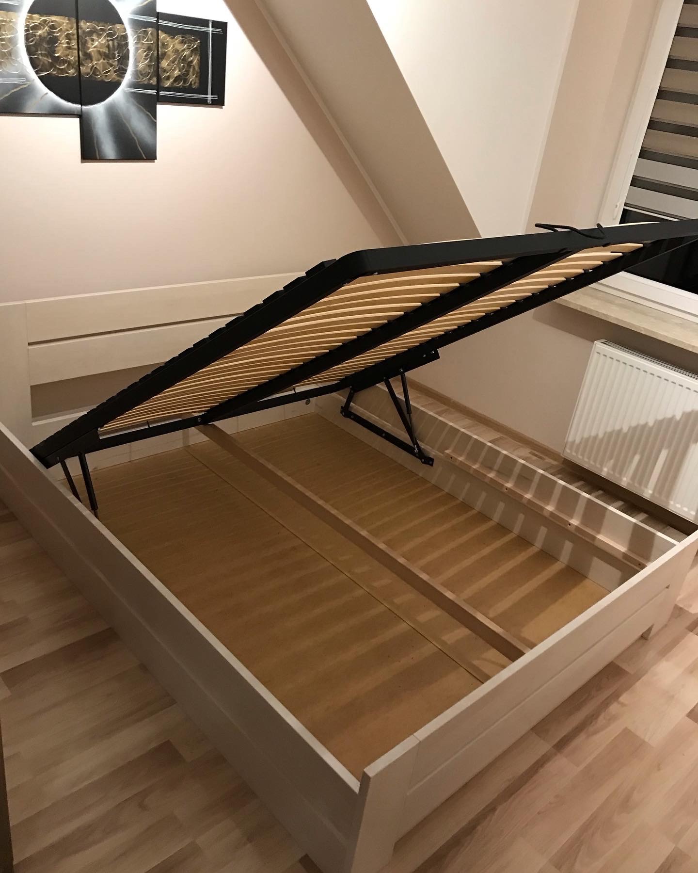 Otwarta rama łóżka z podnoszonym stelażem i pojemnikiem na pościel, widoczne drewniane listwy i mechanizm podnoszenia, na podłodze panele, w tle grzejnik i okno.