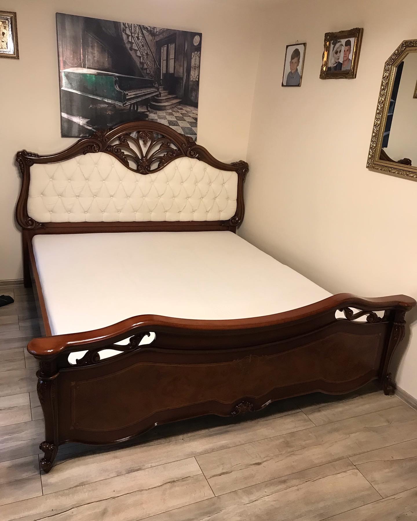 Eleganckie łóżko z ciemnego drewna z kremowym, pikowanym zagłówkiem, na tle ściany z obrazami i lustrem, ustawione na podłodze imitującej drewno.