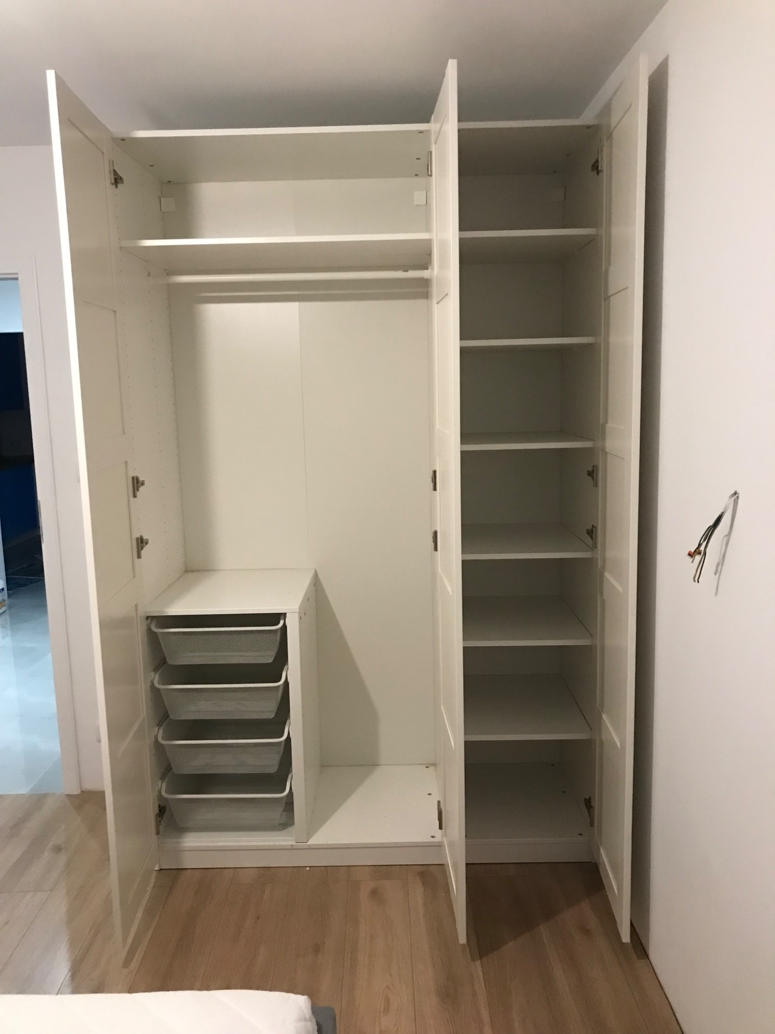 Wnętrze białej szafy z otwartymi drzwiami, widoczne półki, drążek na ubrania i wysuwane kosze, na ścianie wystające przewody elektryczne.