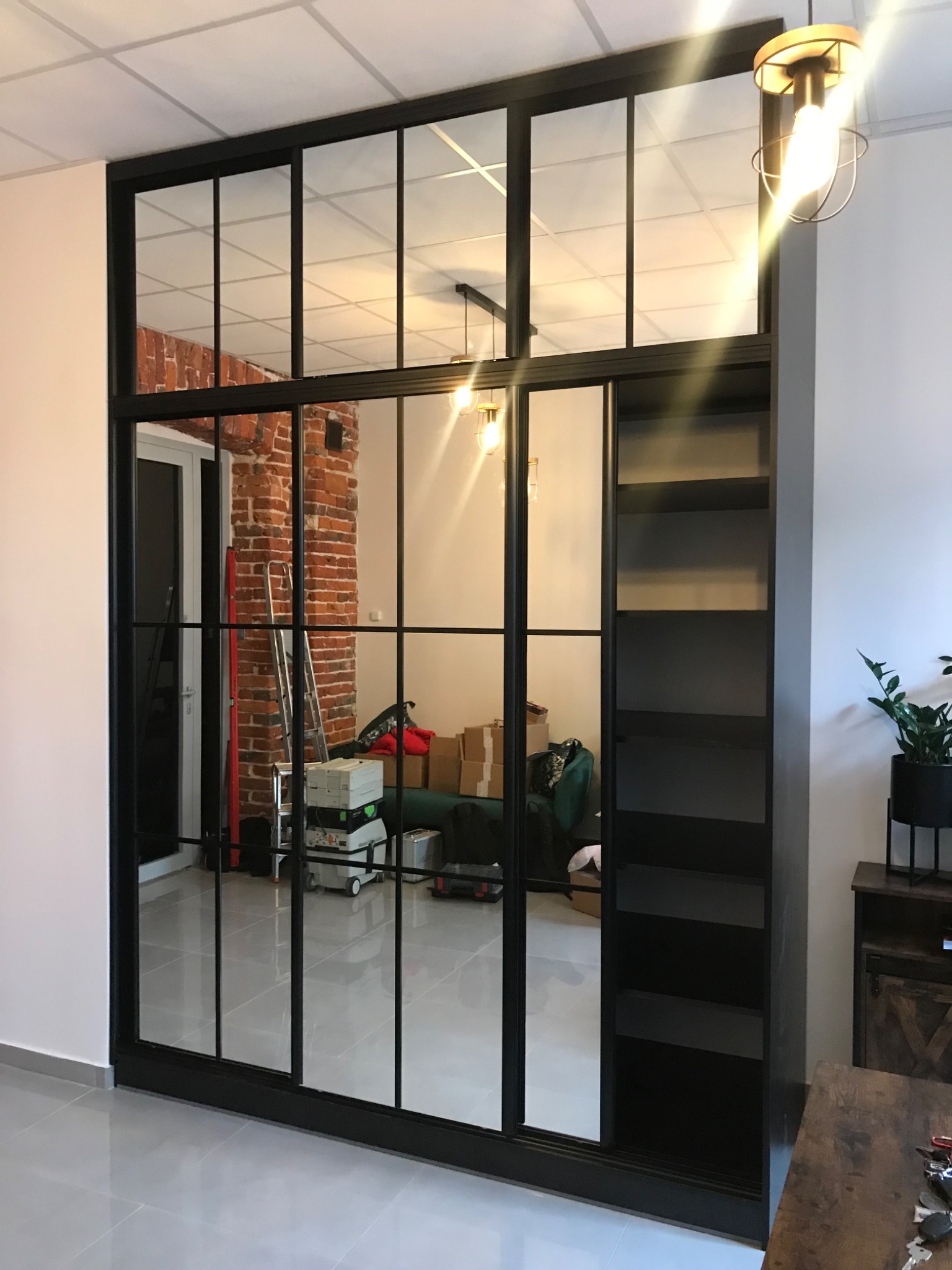 Czarna, przeszklona ścianka działowa w stylu loft z wbudowanym regałem, odbijająca ceglaną ścianę i narzędzia w tle.