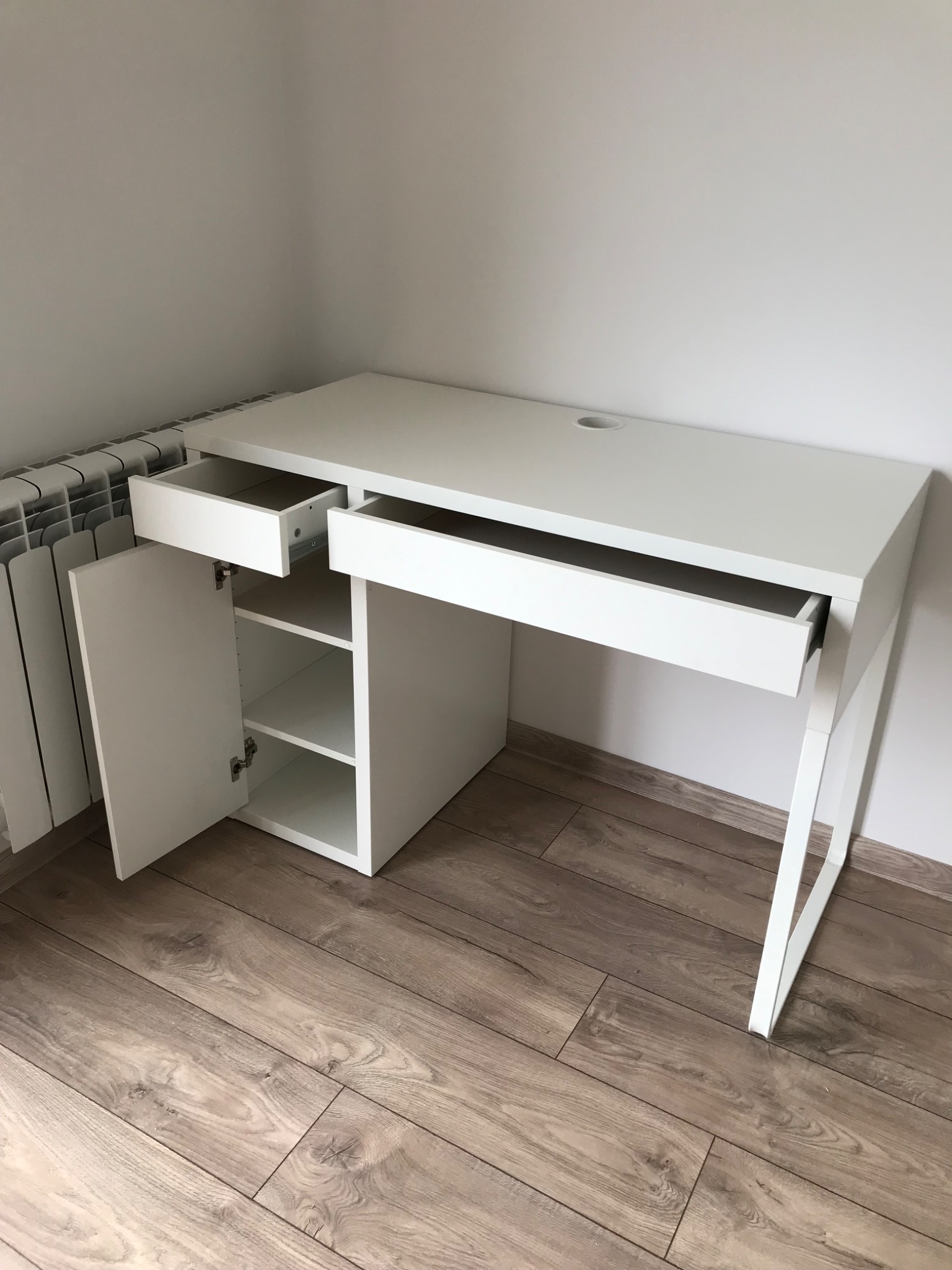 Białe biurko z otwartymi szufladami i szafką, widoczne na tle drewnopodobnej podłogi i grzejnika.