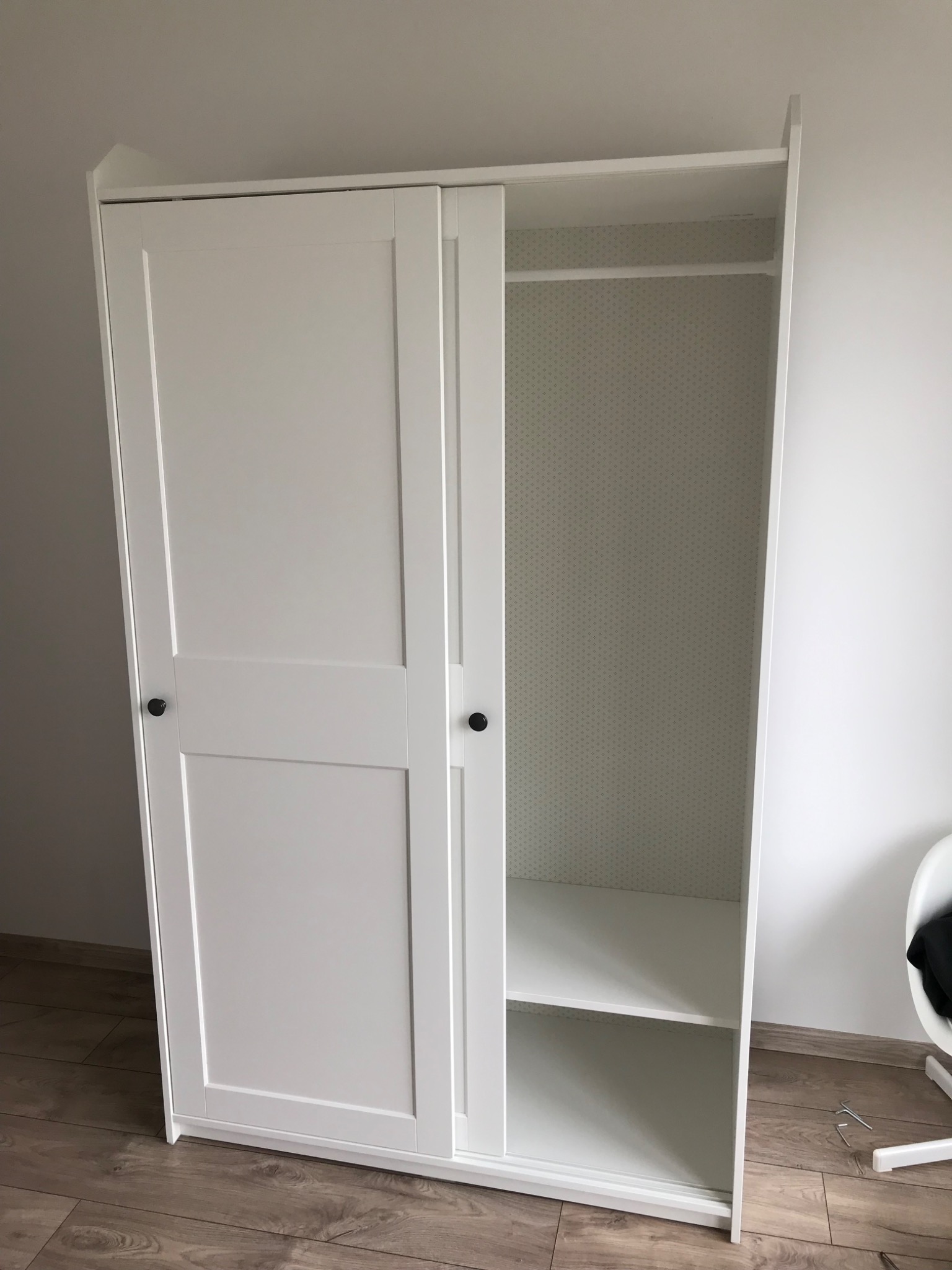 Biała szafa przesuwna z częściowo otwartymi drzwiami, ukazująca półki i drążek na ubrania, ustawiona na podłodze z paneli imitujących drewno, z widocznym narzędziem montażowym leżącym obok.