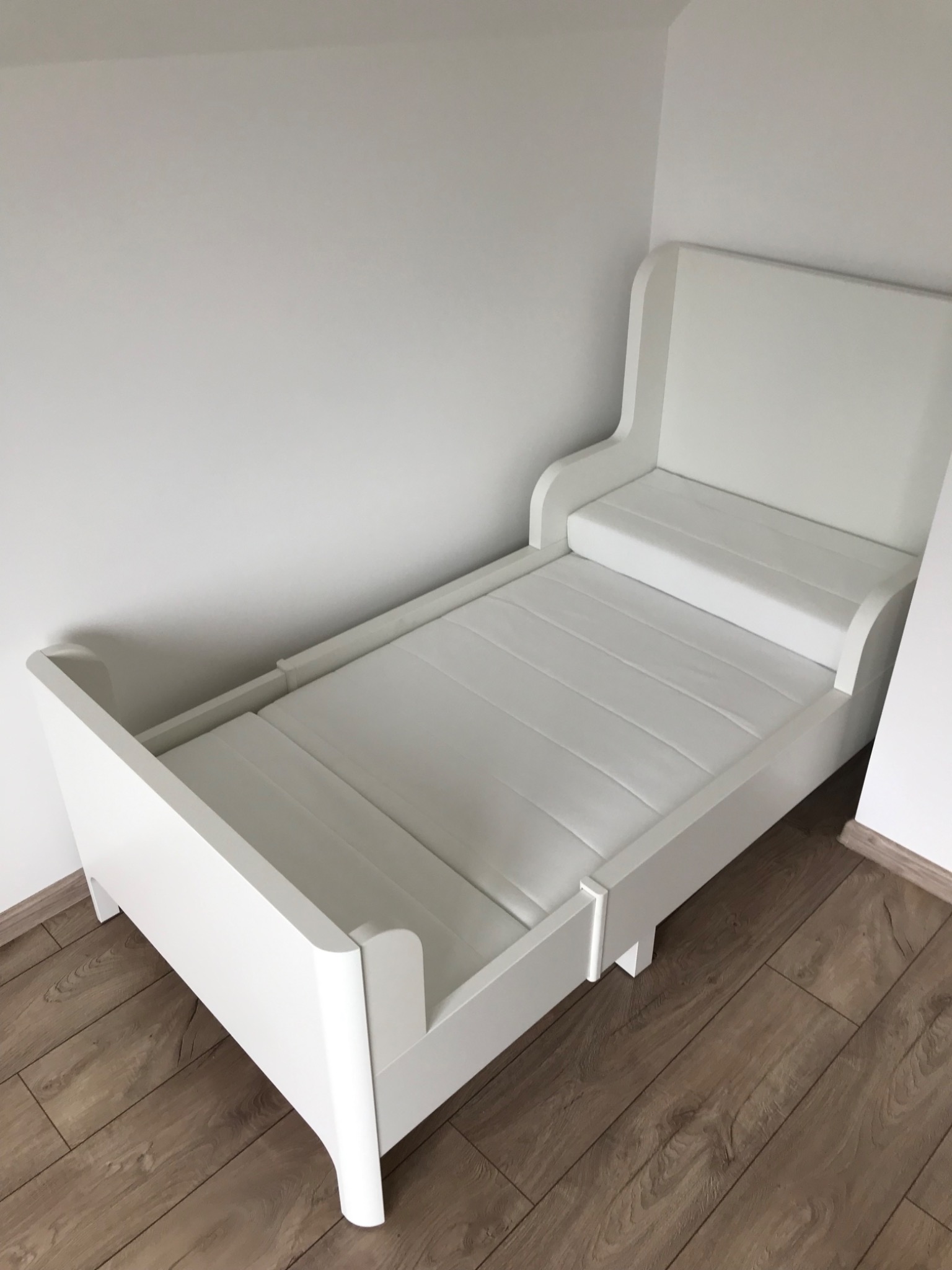 Białe, drewniane łóżeczko dziecięce z materacem, ustawione na panelach podłogowych, sfotografowane w rogu pomieszczenia.