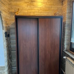 Damian Moskała - Dwudrzwiowa szafa przesuwna z ciemnego drewna, z czarnymi obramowaniami, na tle ściany z dekoracyjnej cegły, oświetlona lampkami w kształcie żółwi.