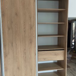 Damian Moskała - Jasna, drewnopodobna szafa przesuwna z otwartą częścią regałową, widoczna szuflada i puste półki, prezentacja konstrukcji i wykończenia.