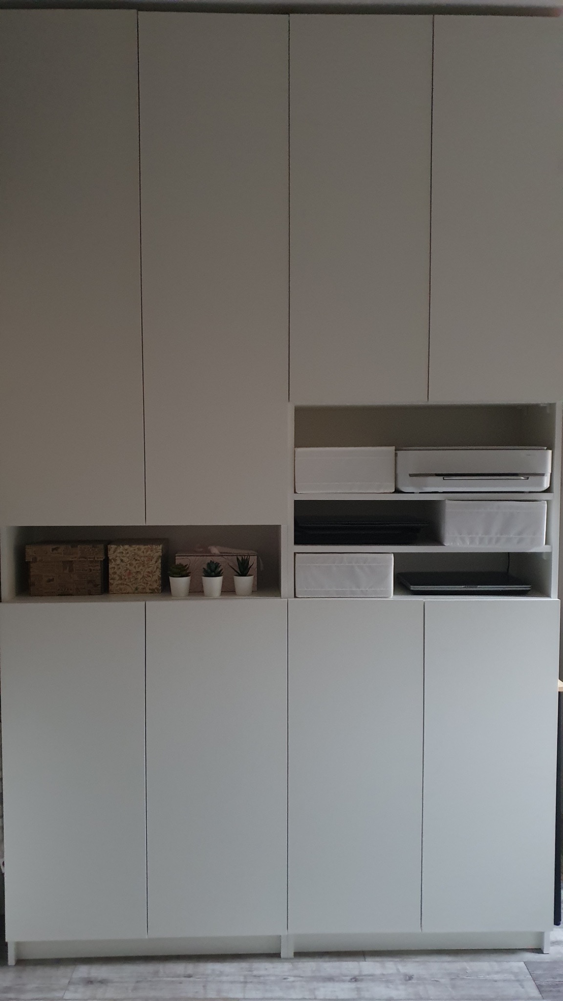 Pełna zabudowa ściany z białymi, gładkimi frontami szafek, z otwartymi półkami zawierającymi dekoracyjne pudełka, sukulenty w doniczkach i białe pojemniki.