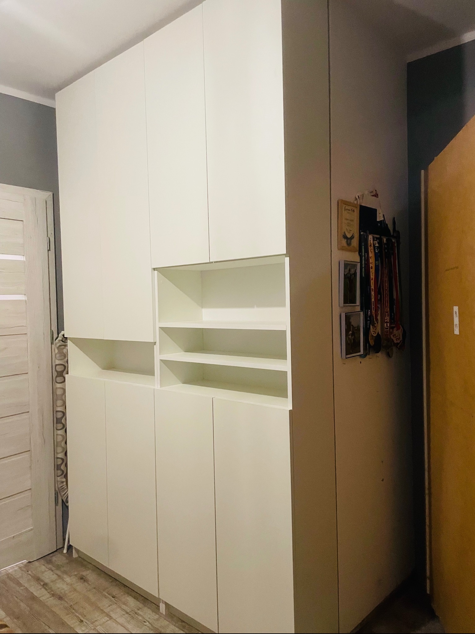 Biała, minimalistyczna szafa z otwartymi półkami w górnej części, widoczne medale wiszące na ścianie obok