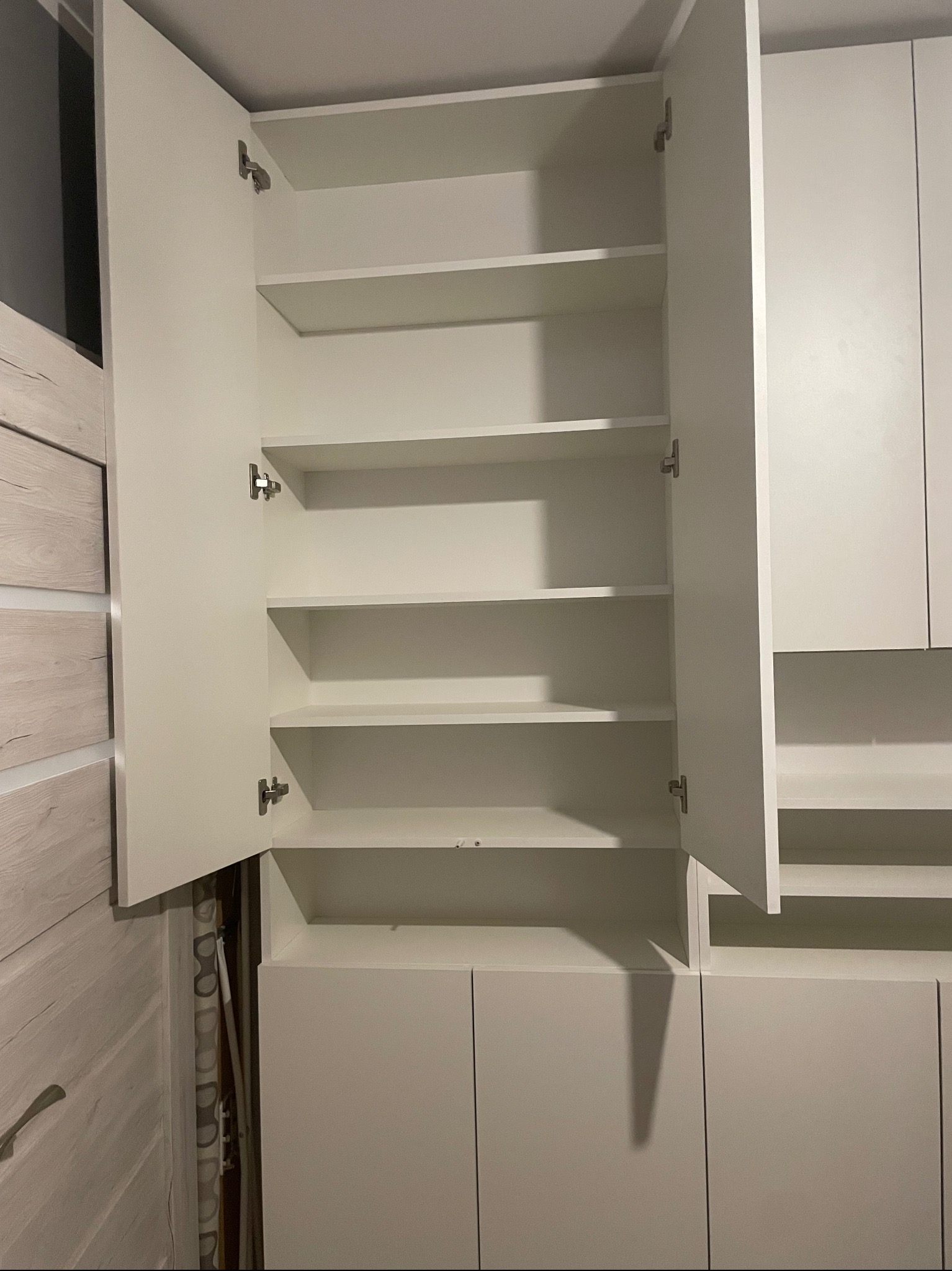 Biała szafa z otwartymi drzwiami, widoczne półki i zawiasy, ściana z jasnego drewna w tle.