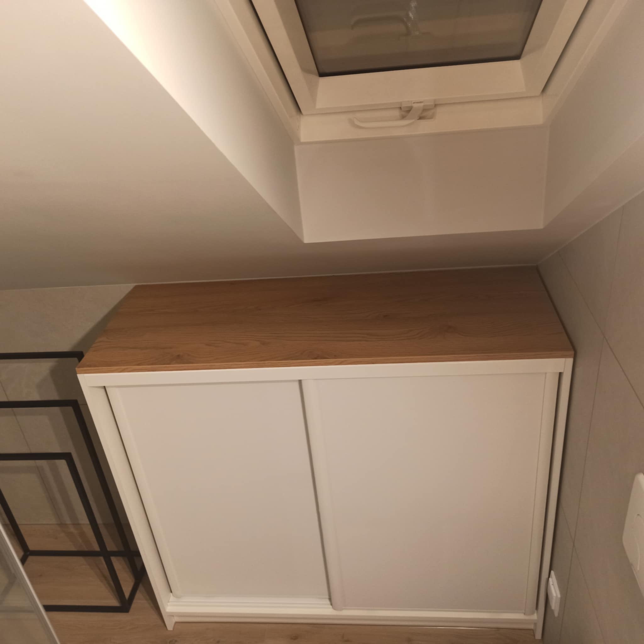 Biała szafa z przesuwnymi drzwiami i drewnianym blatem, umieszczona pod oknem dachowym na poddaszu.