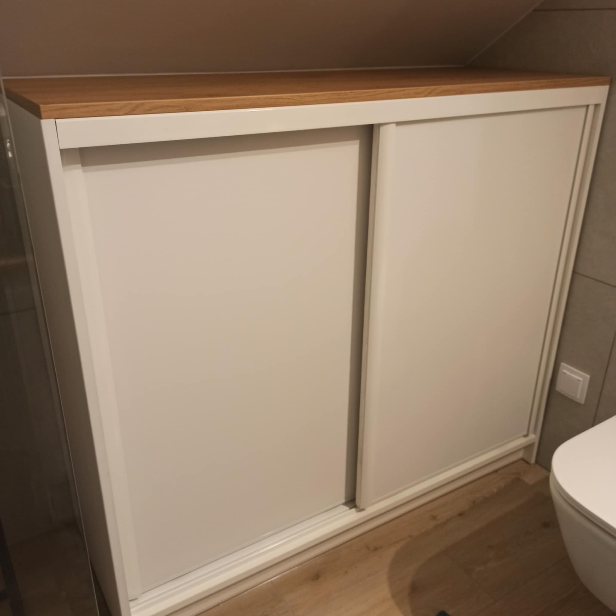 Biała szafa z przesuwnymi drzwiami i drewnianym blatem w przestrzeni ze skosem, widoczny fragment toalety.