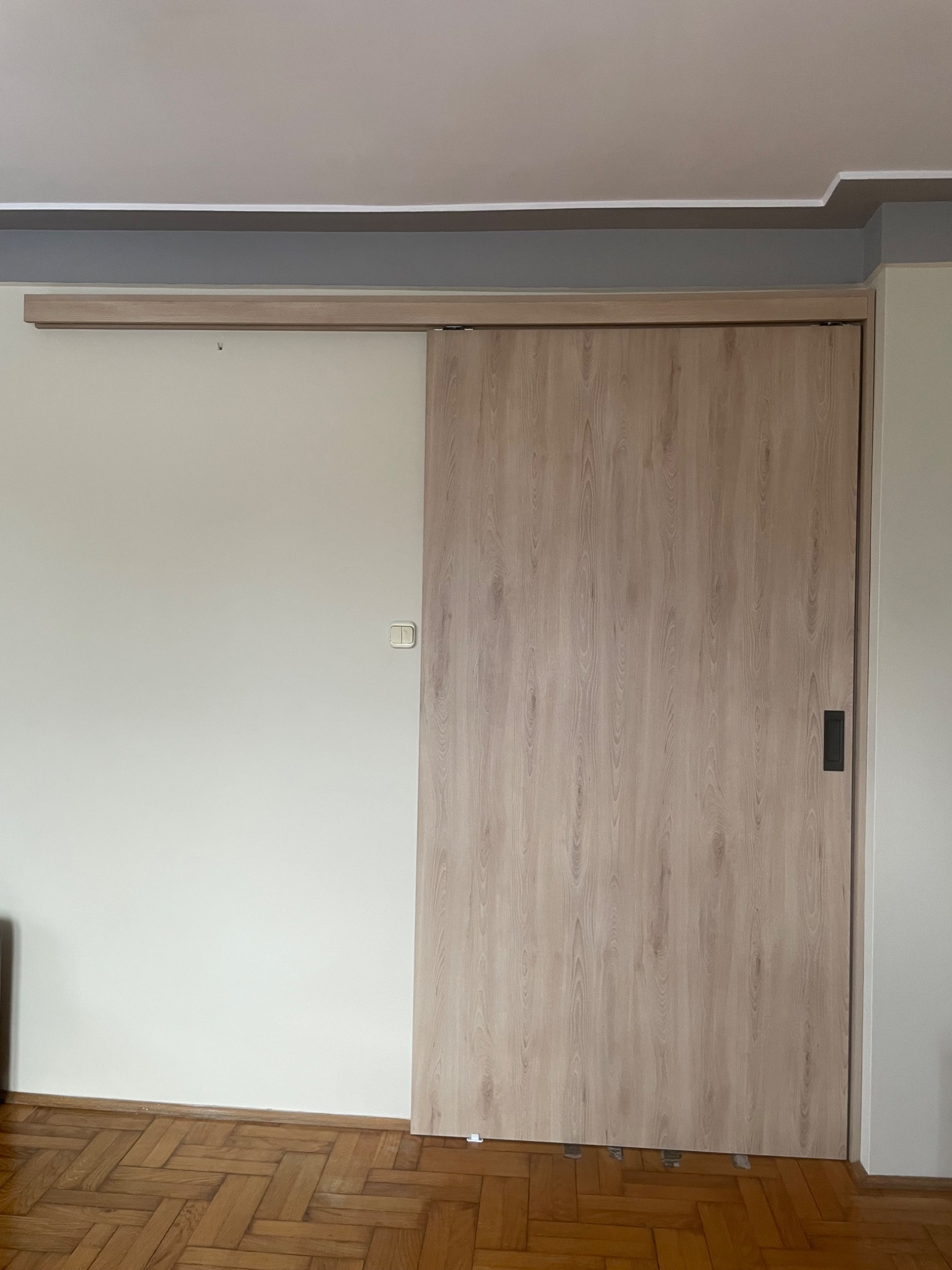 Jasna, drewnopodobna przesuwna szafa wnękowa w Krakowie, częściowo zasłaniająca ścianę z włącznikiem, na tle parkietu ułożonego w jodełkę.