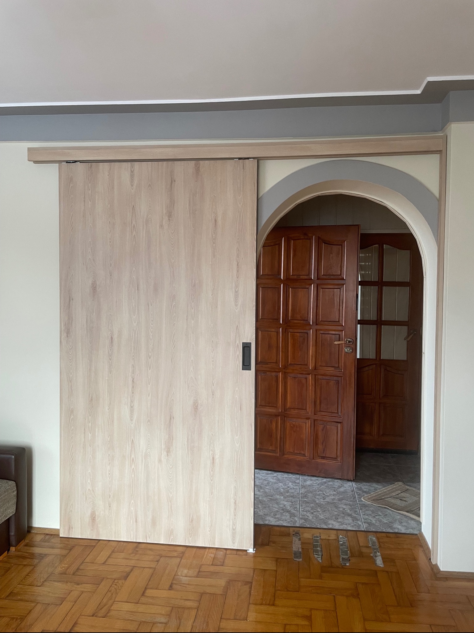 Jasna, drewnopodobna przesuwana szafa z czarnym uchwytem, częściowo zasłaniająca wejście z łukowym nadprożem, w tle widoczne drewniane drzwi z szybkami.