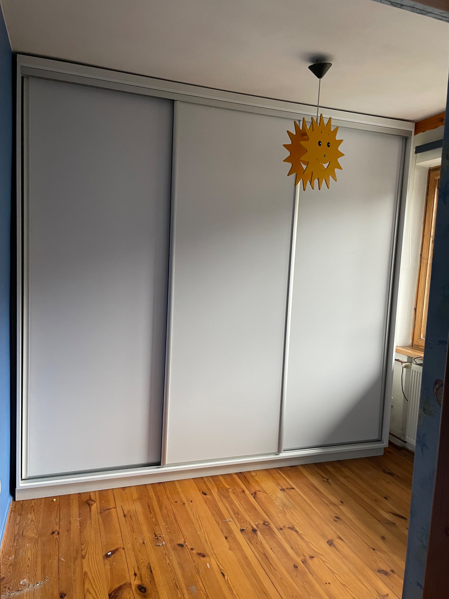 Trzydrzwiowa szafa przesuwna z białymi frontami, widok na drewnianą podłogę i lampę w kształcie słońca.