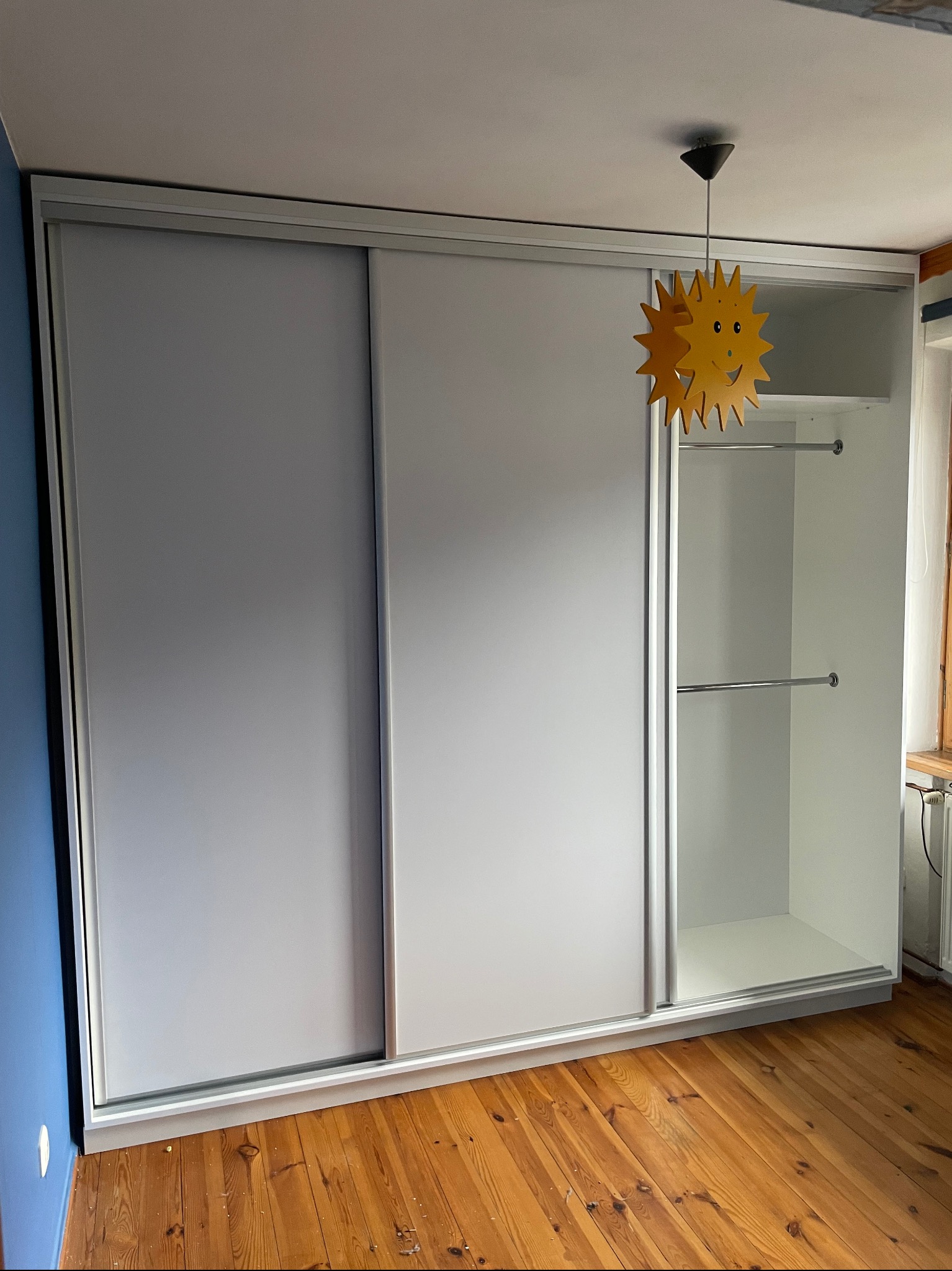 Biała szafa wnękowa z przesuwnymi drzwiami, widoczne wnętrze z drążkami na ubrania, oświetlona lampą w kształcie słońca, drewniana podłoga.