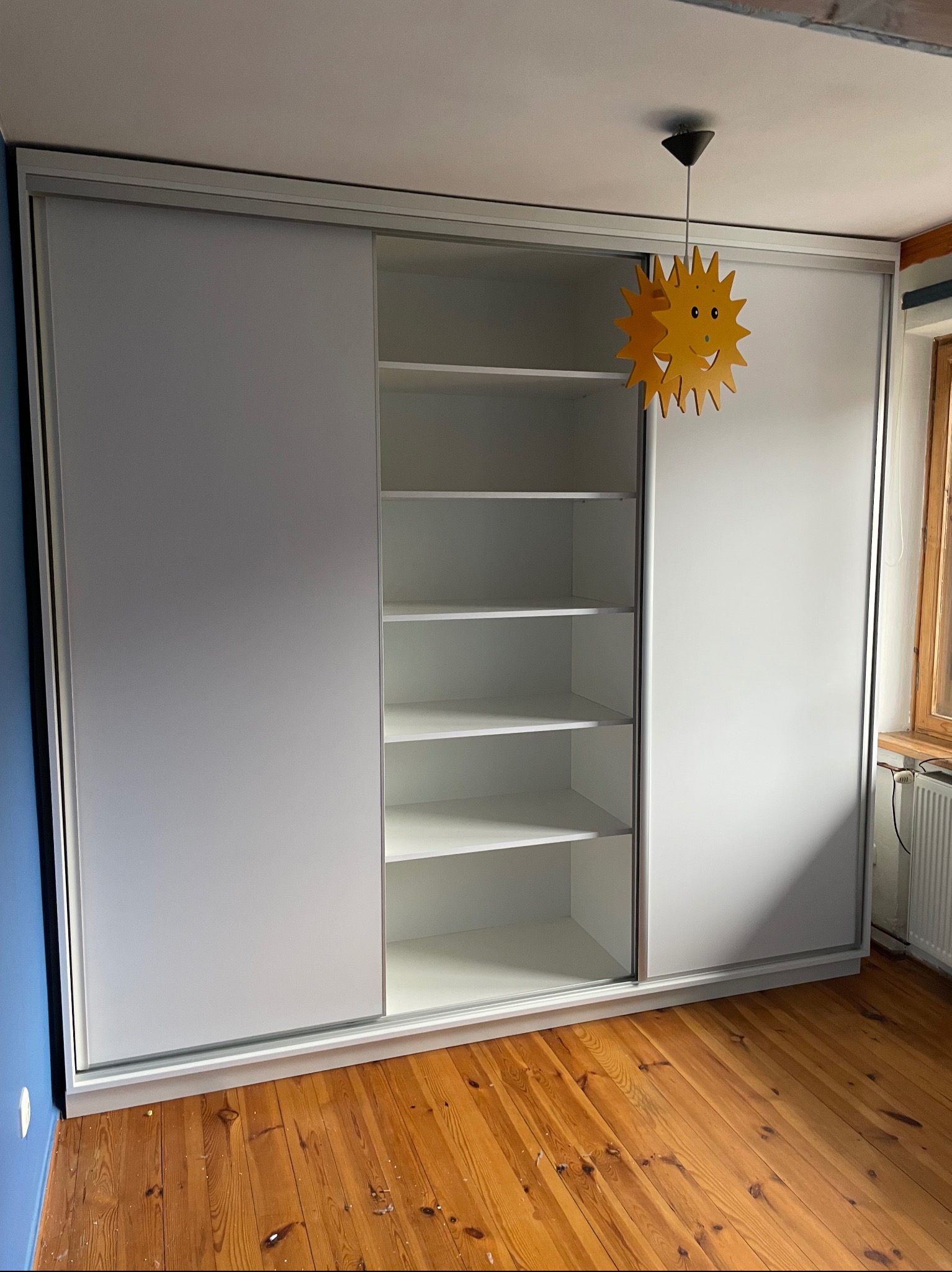 Biała szafa przesuwna z otwartą środkową częścią, ukazującą puste półki, na tle drewnianej podłogi i niebieskiej ściany, z wiszącą lampą w kształcie słońca.