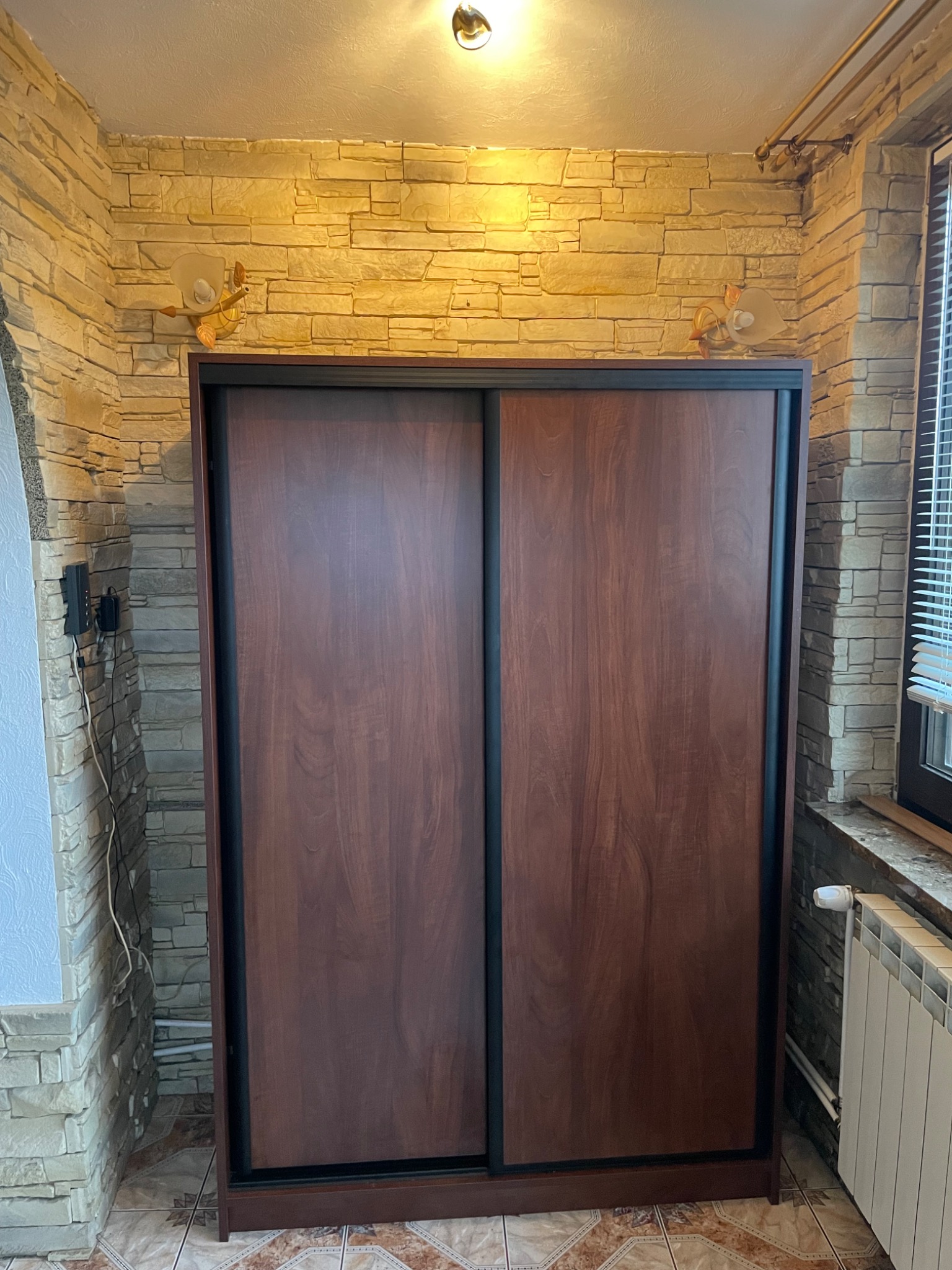 Dwudrzwiowa szafa przesuwna z ciemnego drewna, z czarnymi obramowaniami, na tle ściany z dekoracyjnej cegły, oświetlona lampkami w kształcie żółwi.