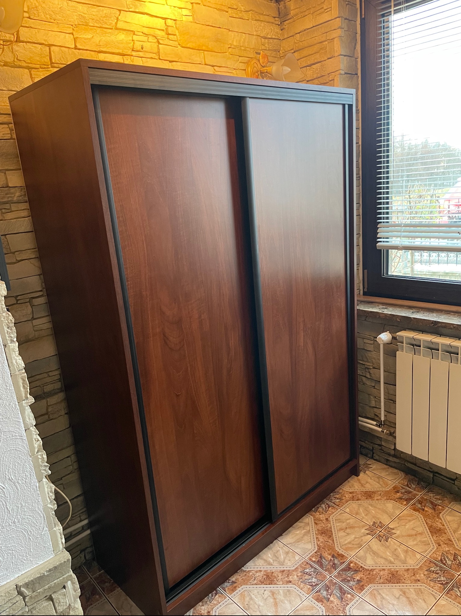 Wysoka, brązowa szafa z przesuwnymi drzwiami i czarnymi obramowaniami, ustawiona w rogu pokoju z kamienną ścianą i wzorzystą podłogą z płytek, obok okna z żaluzjami i grzejnika.