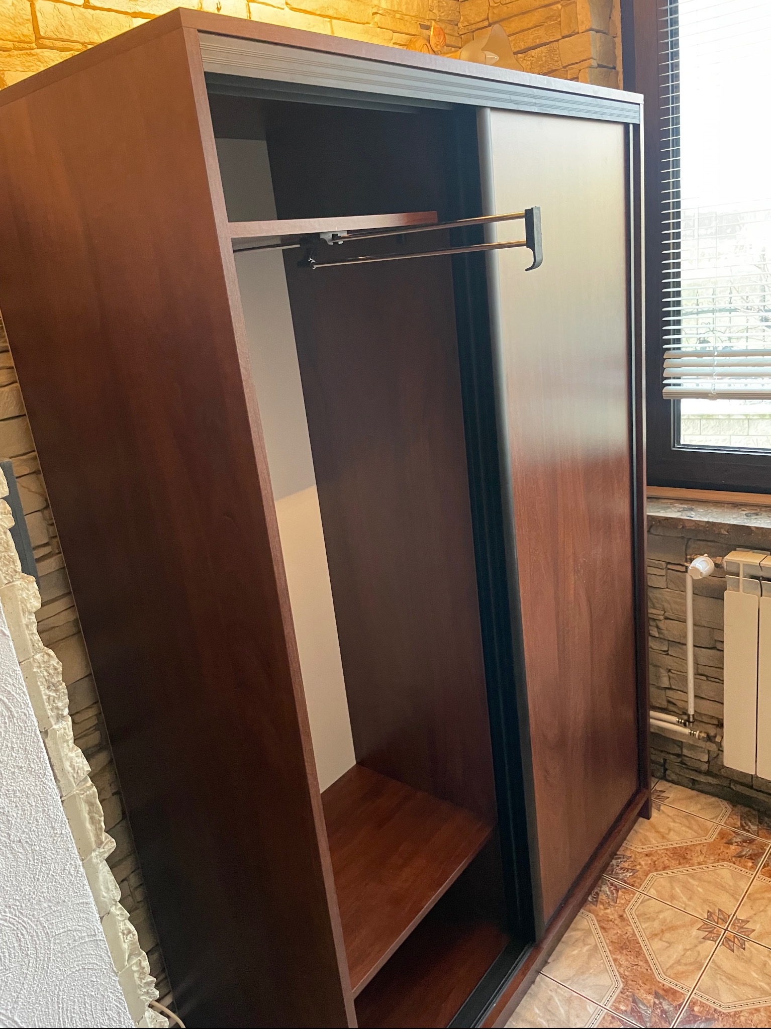 Ciemnobrązowa szafa z przesuwnymi drzwiami i otwartą częścią z półkami oraz drążkiem na ubrania. Widoczna faktura drewna i czarne obramowanie drzwi. W tle okno z żaluzjami i grzejnik.