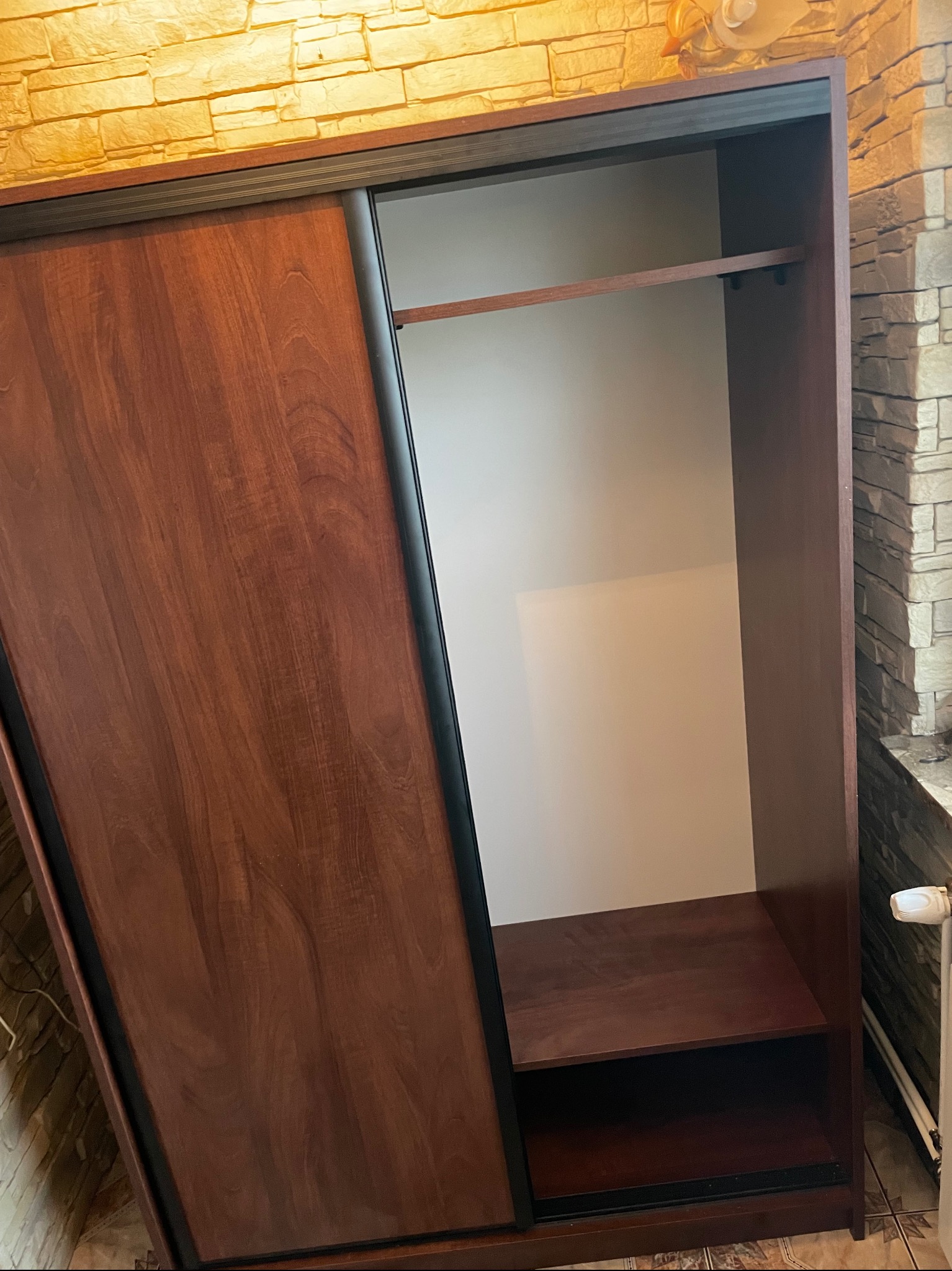 Częściowo otwarta, brązowa szafa z przesuwnymi drzwiami, widoczna półka i drążek na wieszaki, tło stanowią dekoracyjne ściany z imitacji kamienia.