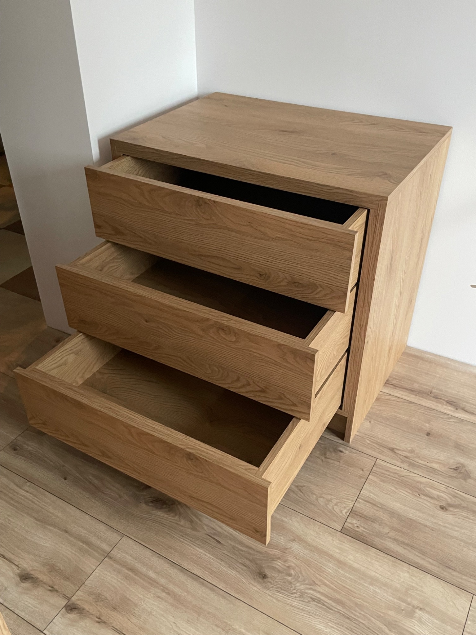 Komoda z trzema szufladami z jasnego drewna dębowego, z otwartymi szufladami, ukazująca minimalistyczny design i dopasowanie do podłogi w kolorze drewna.
