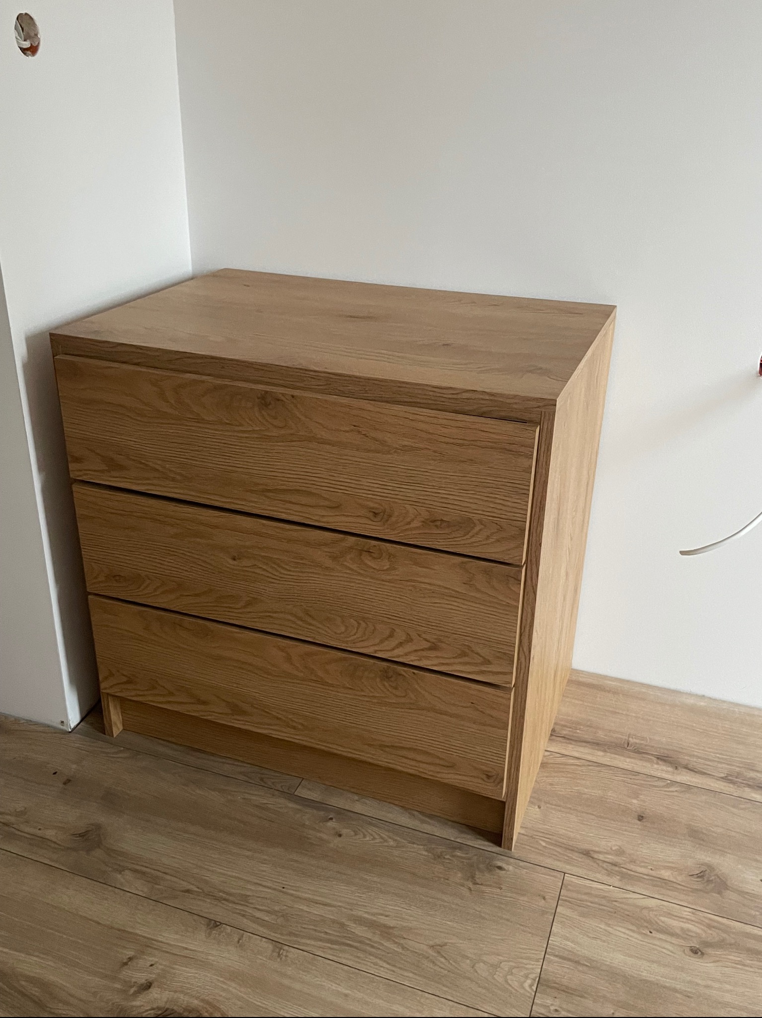 Trzy-szufladowa komoda z jasnego drewna dębowego, ustawiona na drewnianej podłodze w rogu pokoju, widoczne niedokończone prace wykończeniowe na ścianie.